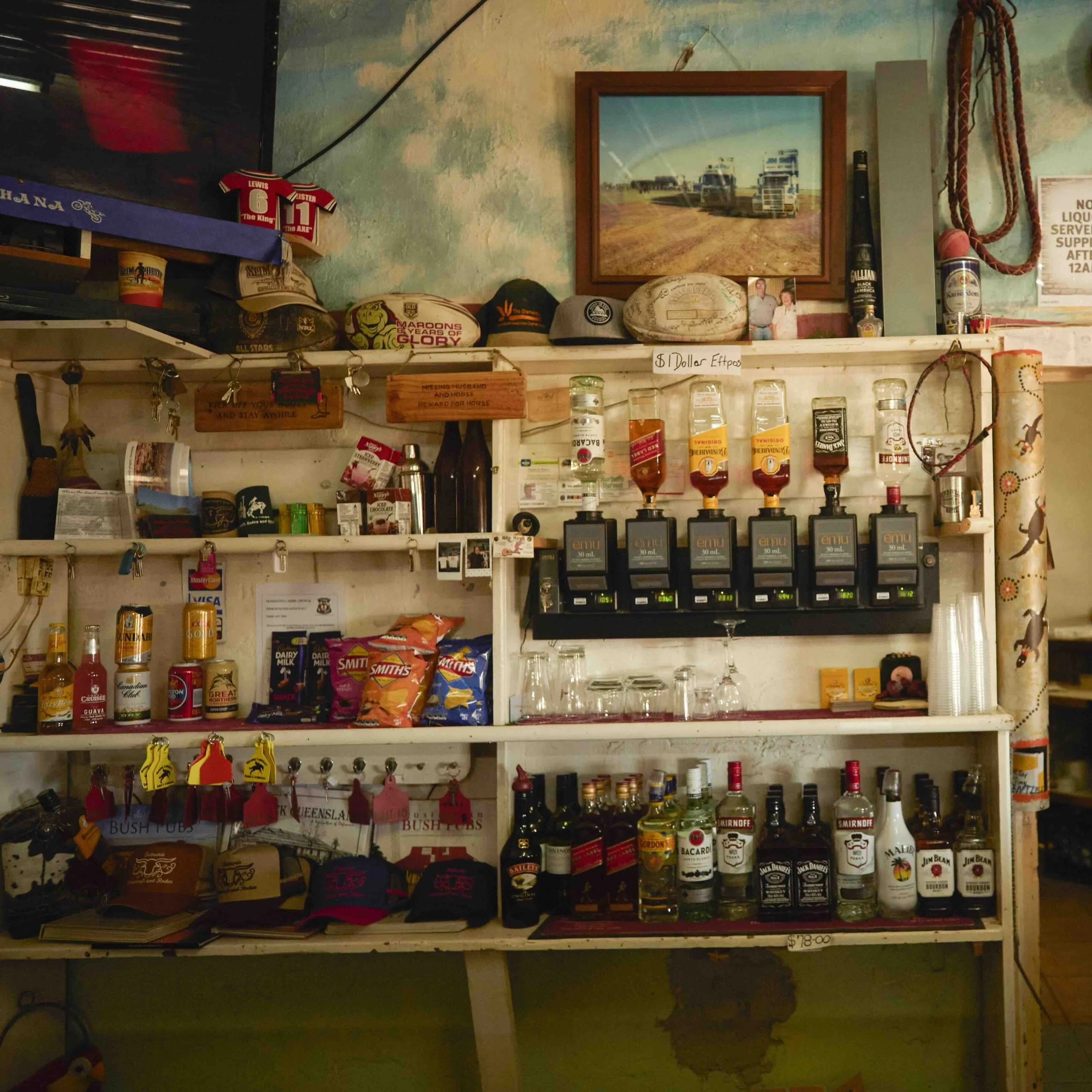 A rustic outback bar interior with wooden shelves stocked with liquor bottles, spirit dispensers, and snacks. The shelves also display various memorabilia, including trucker caps, sports merchandise, and a framed photo of road trains in a dusty landscape.