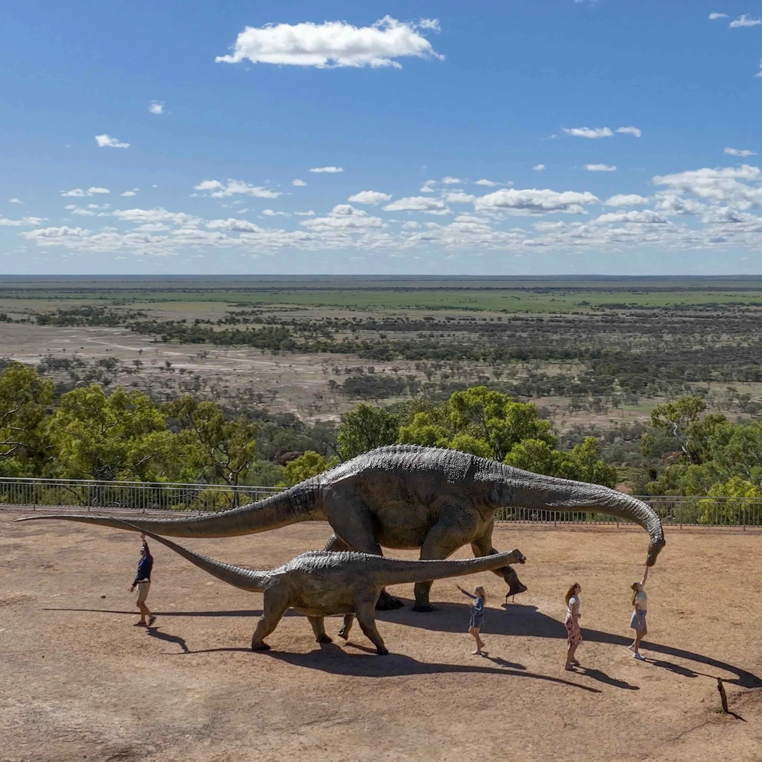 A scenic outdoor exhibit features two large dinosaur sculptures, one adult and one juvenile, against a vast outback landscape with green vegetation and open plains under a bright blue sky. Visitors, including children, interact with the sculptures, reaching out to touch the dinosaurs.