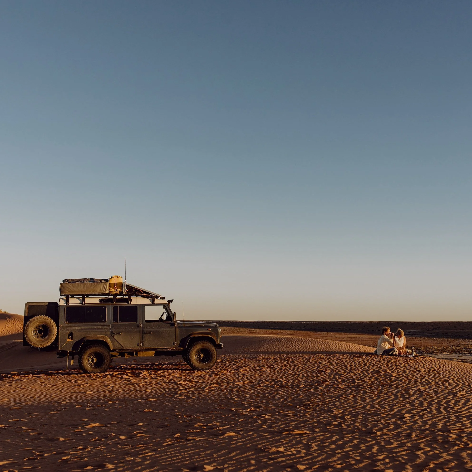 A rugged off-road vehicle with a roof rack and camping gear is parked on a vast, sandy desert landscape under a clear sky during sunset. Nearby, two people sit on the sand, enjoying the peaceful surroundings.