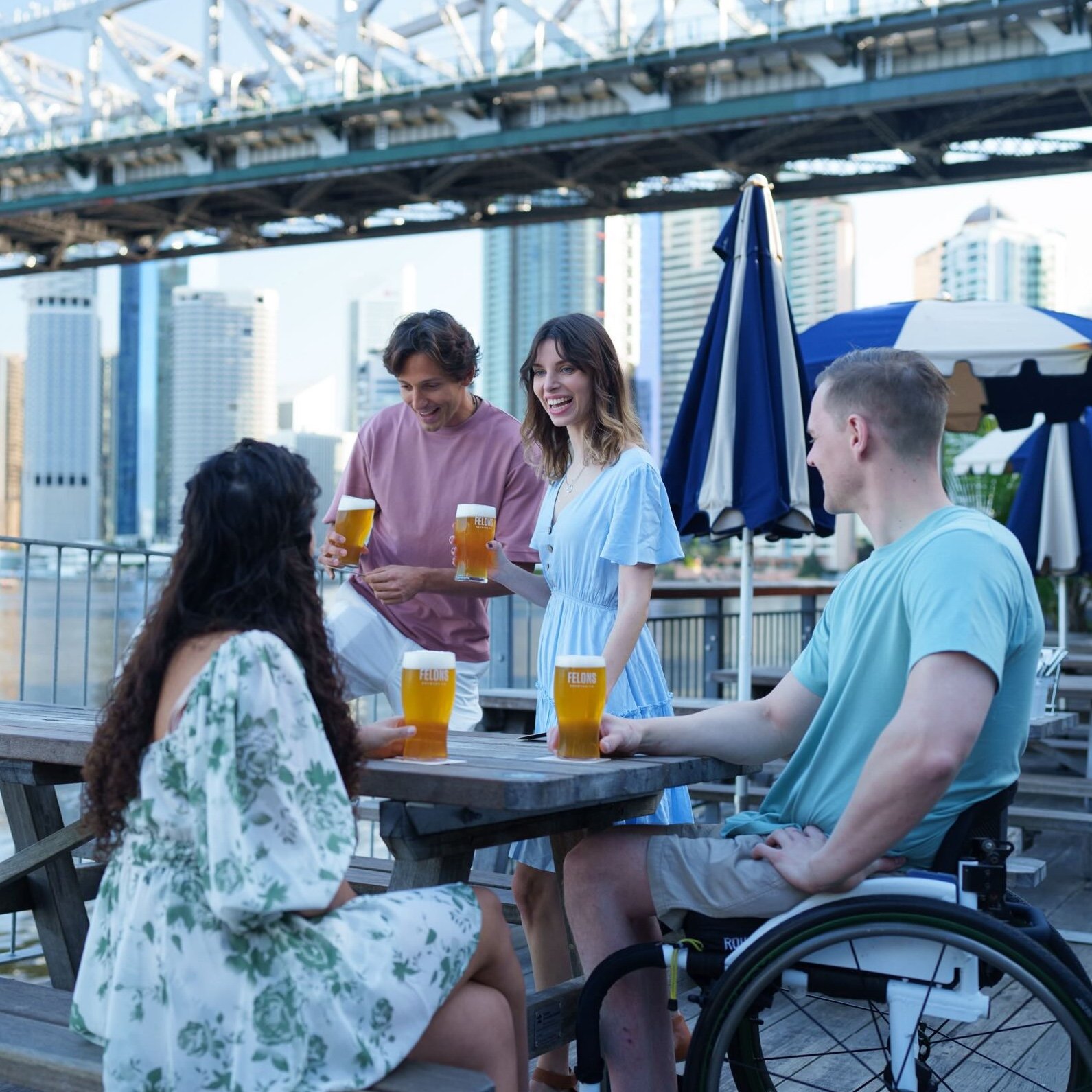 A cheerful group of friends enjoying drinks at an outdoor venue, with a bridge and city skyline in the background.