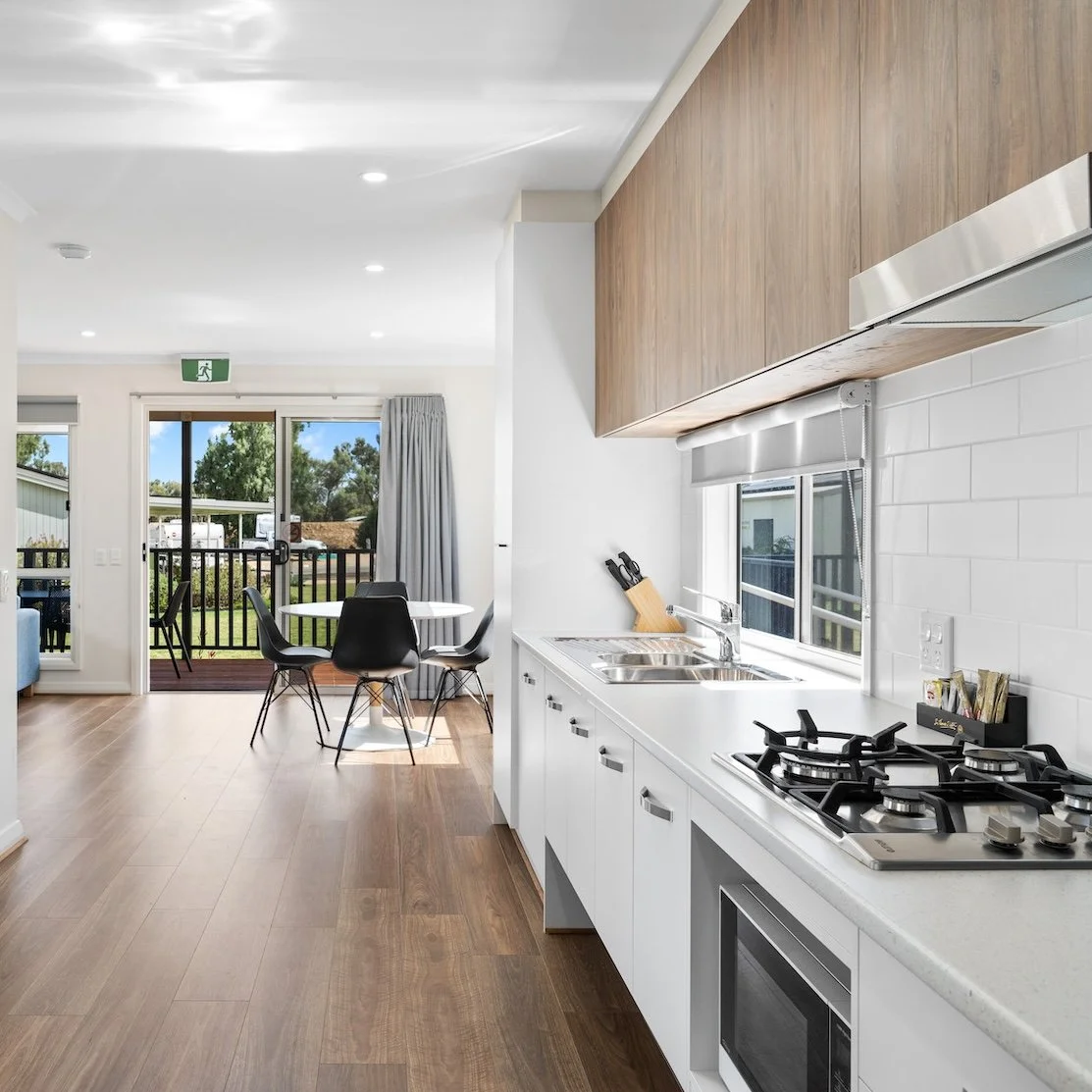 Modern holiday cabin interior featuring a sleek kitchen with white cabinetry, stainless steel appliances, and wooden upper cabinets. The space opens to a small dining area with black chairs and a round table, and a view of the outside patio through sliding glass doors.