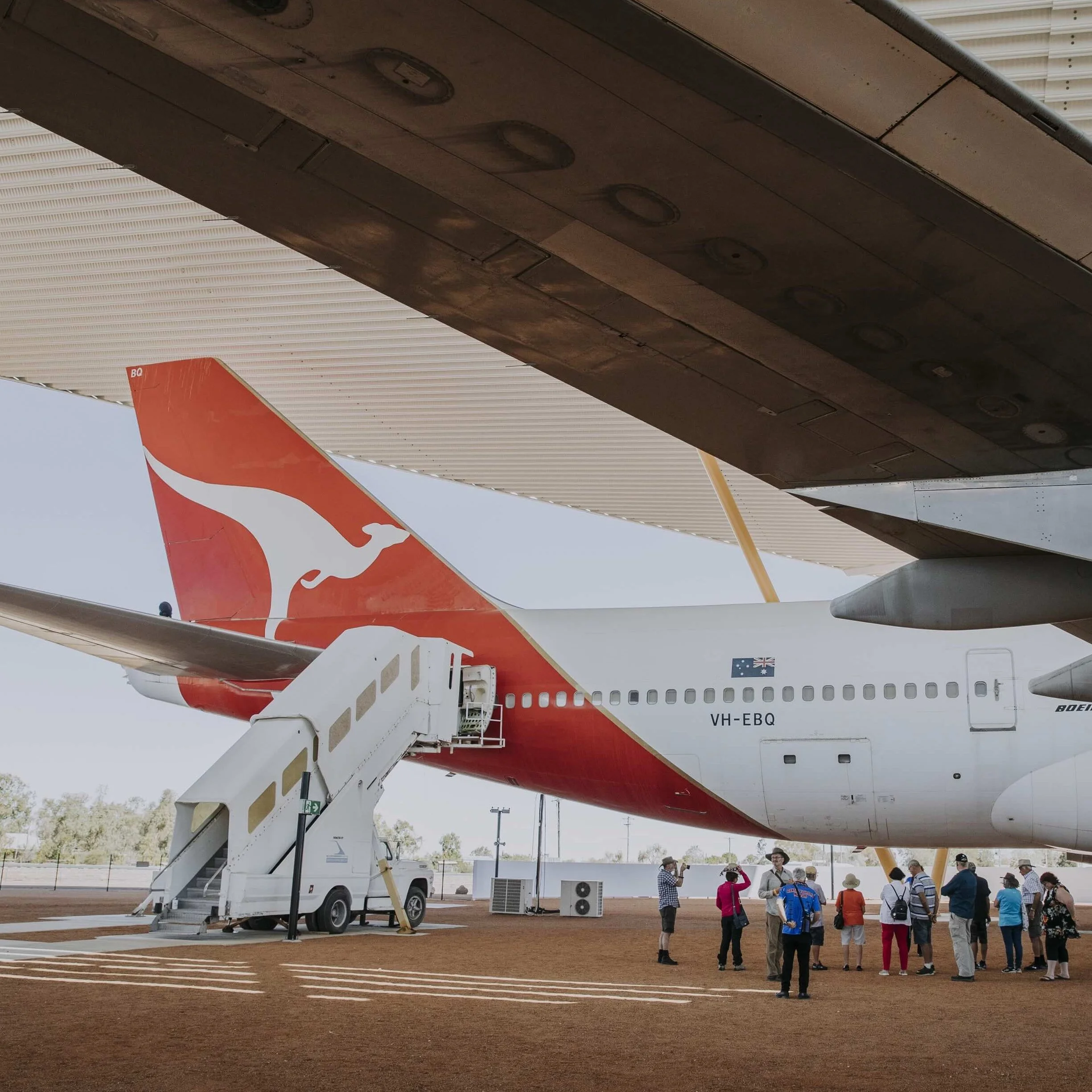 A retired Qantas Boeing 747 aircraft is displayed under a large shelter, with a group of visitors gathered around it. The plane’s red and white tail fin features the iconic kangaroo logo. 