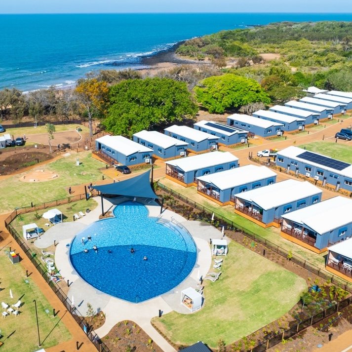 Aerial view of a coastal holiday park featuring rows of modern cabins, a large resort-style swimming pool with a shaded area, and well-maintained green lawns.
