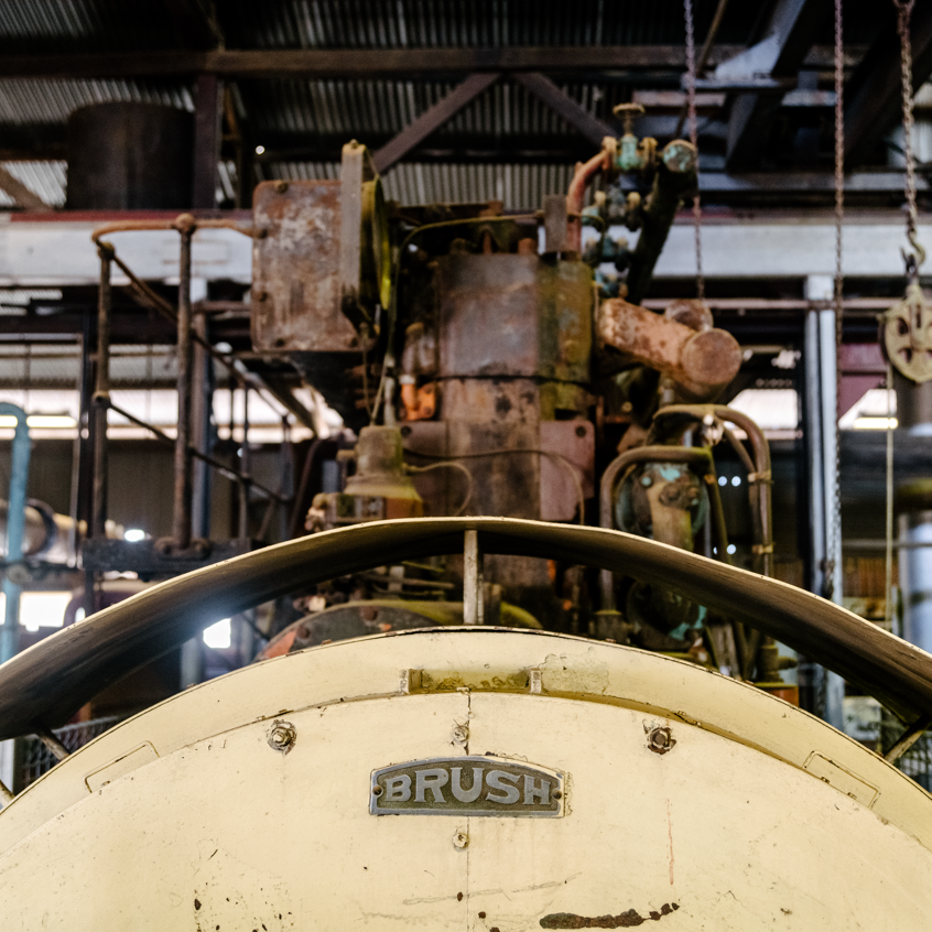A close-up view of an old industrial machine inside a historic powerhouse museum. The foreground features a large, curved metal structure labeled ‘BRUSH,’ while the background reveals a rusted, mechanical assembly with pipes, gears, and levers.