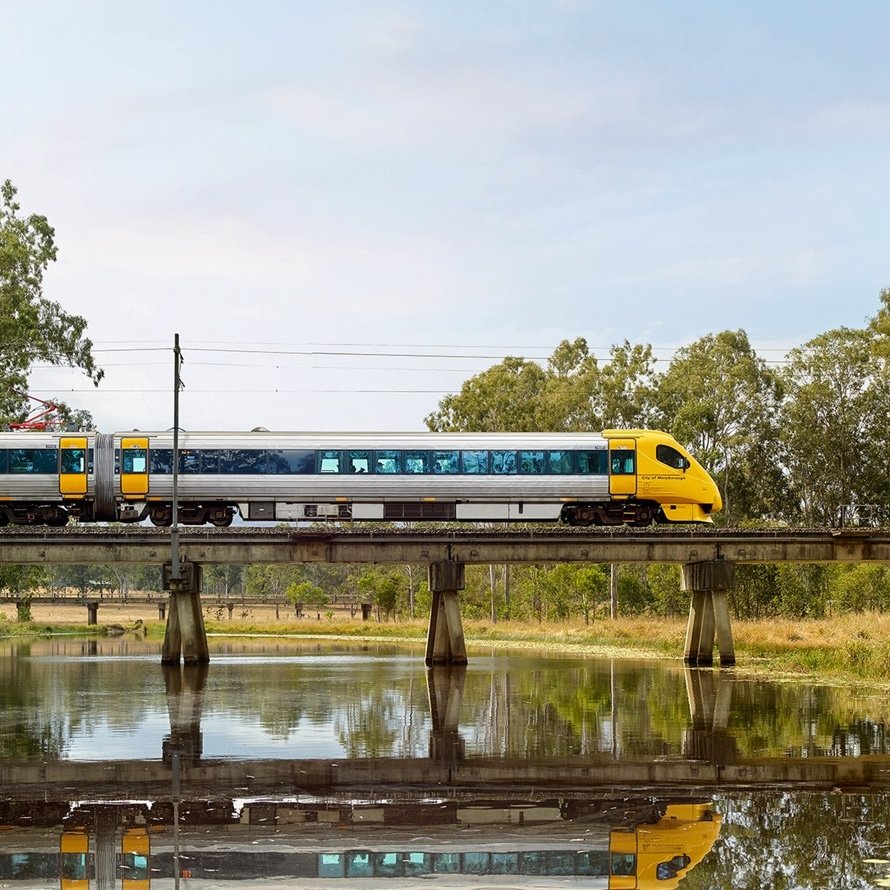 A modern yellow and silver passenger train crossing a bridge over a calm body of water, with trees and greenery in the background. The reflection of the train is visible in the water below, under a lightly cloudy sky.
