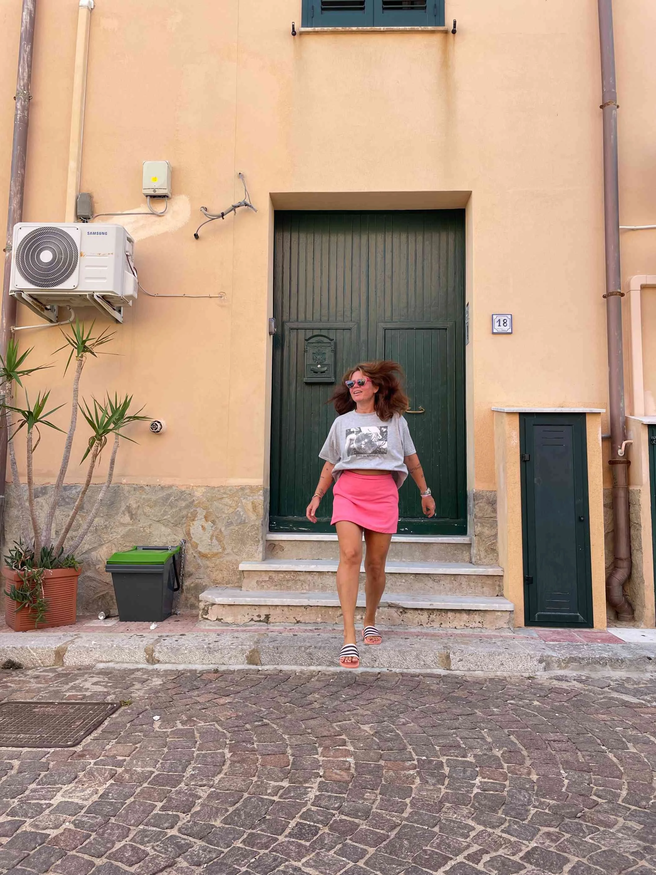 Woman with long hair, wearing sunglasses, a gray T-shirt with a graphic print, a pink skirt, and striped slides, walking down stone steps outside a beige building with a green door.