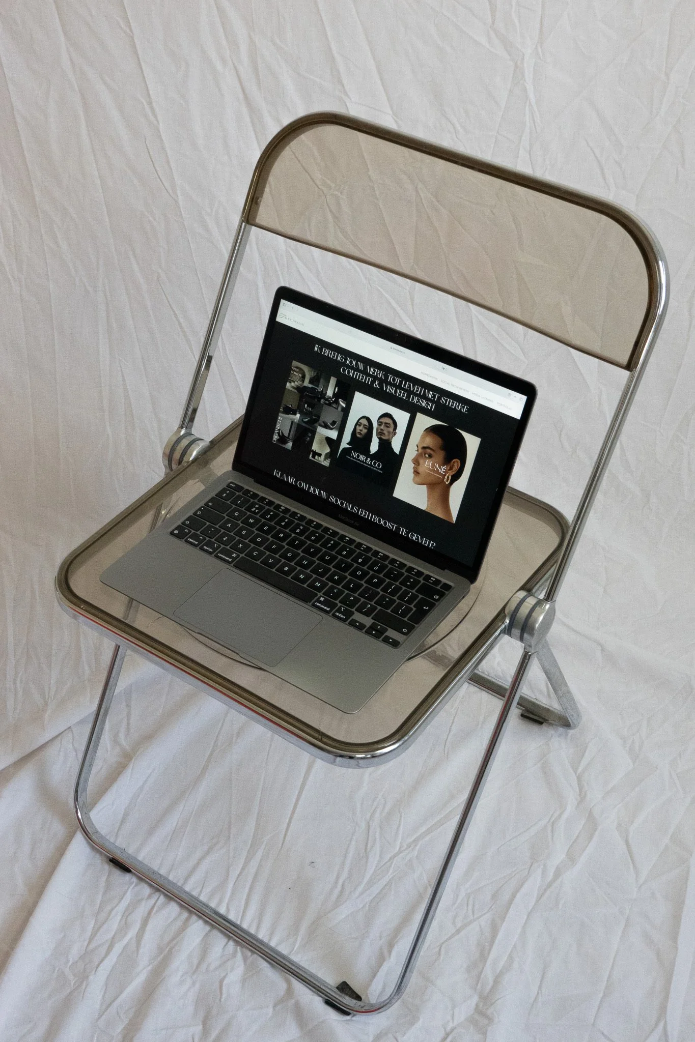Open laptop displaying a fashion website on a chrome-framed glass chair atop a white bed