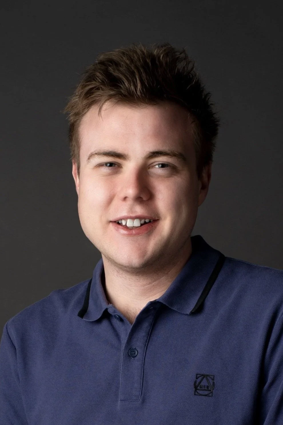 A man smiling in a dark polo shirt with a logo, neutral background.