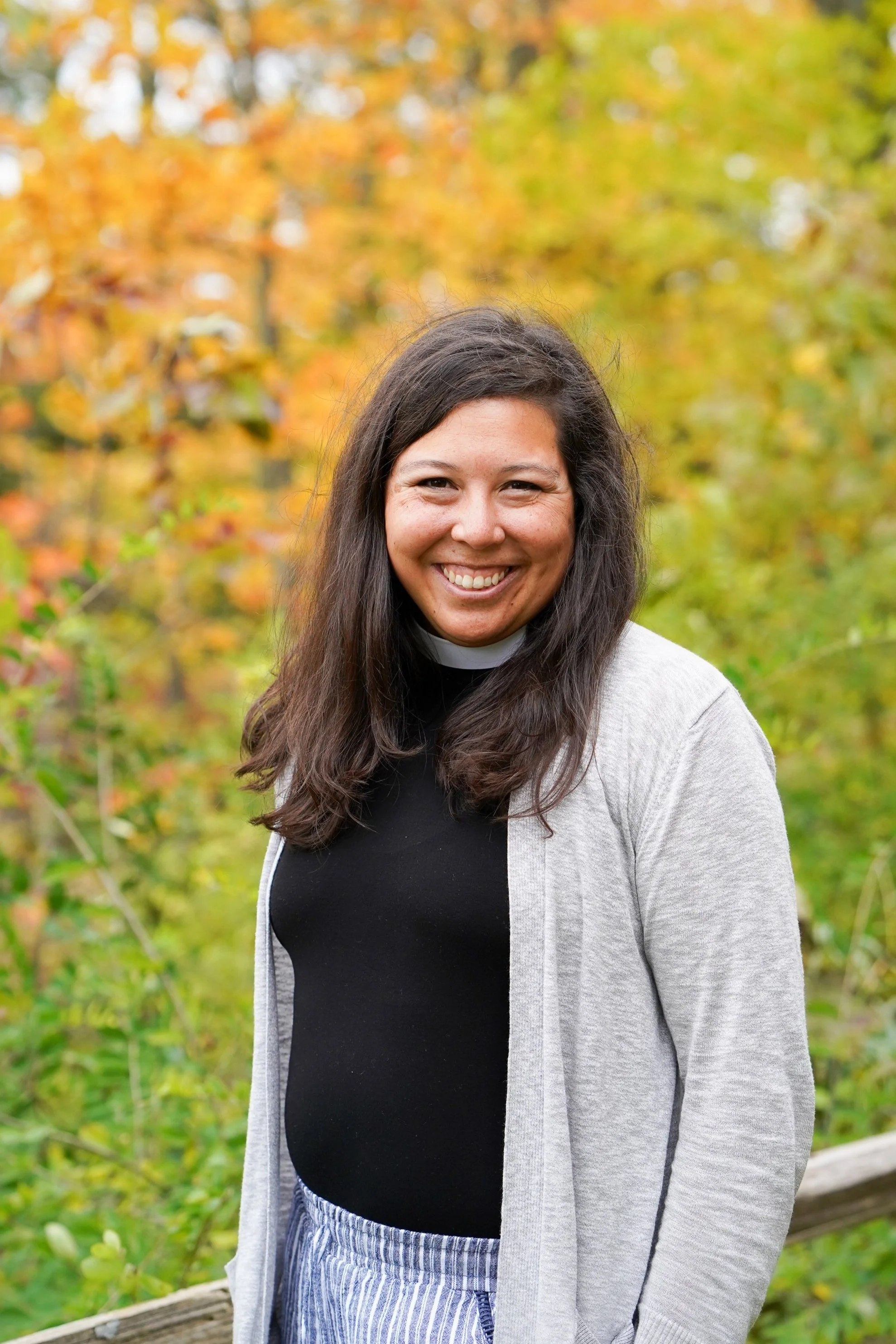 A smiling woman with long dark hair standing outdoors in front of fall-colored trees.