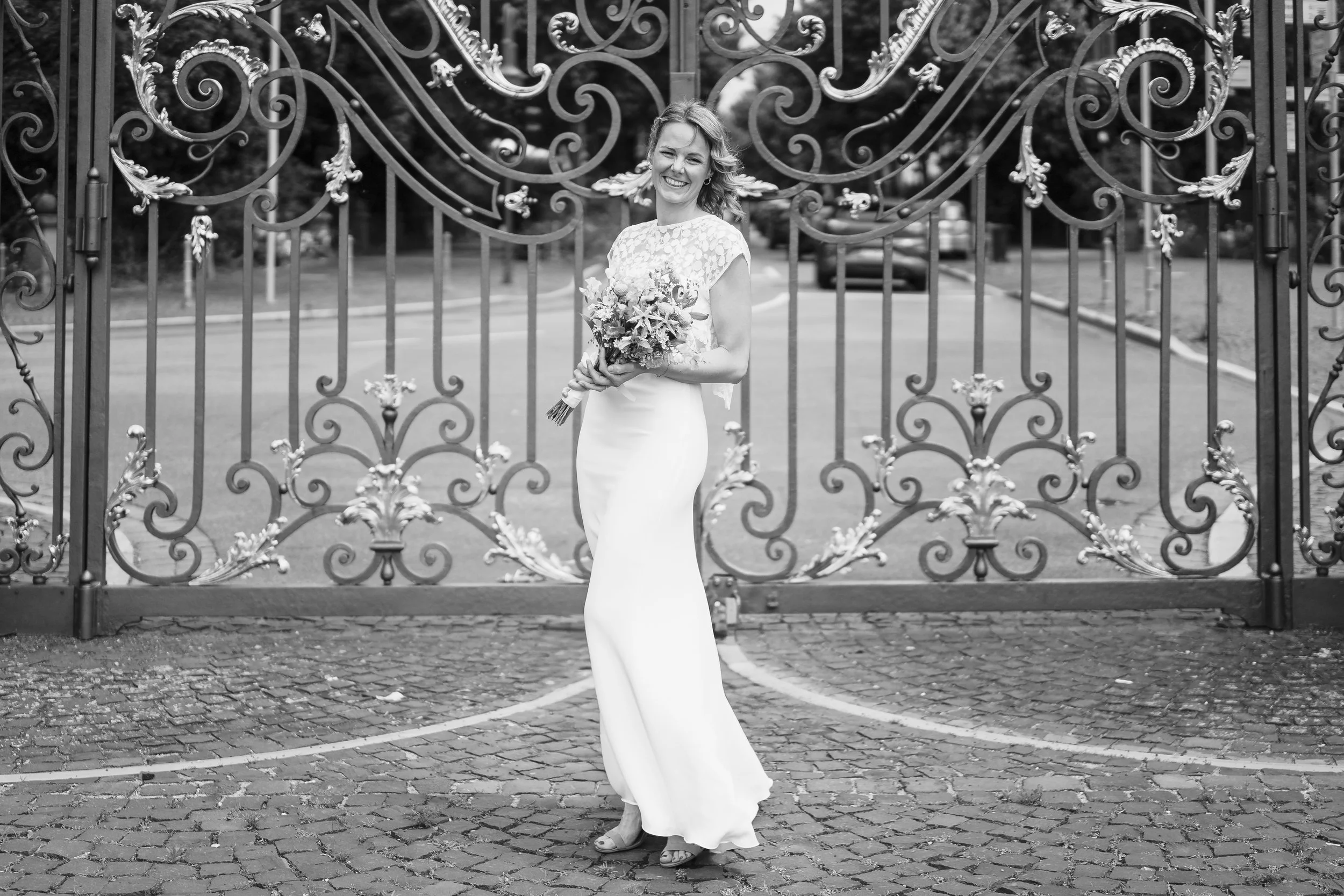 Eine lachende Frau in einem langen weißen Brautkleid, die einen Blumenstrauß hält, steht vor einem kunstvoll verzierten Tor auf gepflastertem Boden. Brautpaarshooting, Paarshooting, Hochzeitsfotos Schloss Philippsruhe Hanau. Standesamt, Frankfurt.