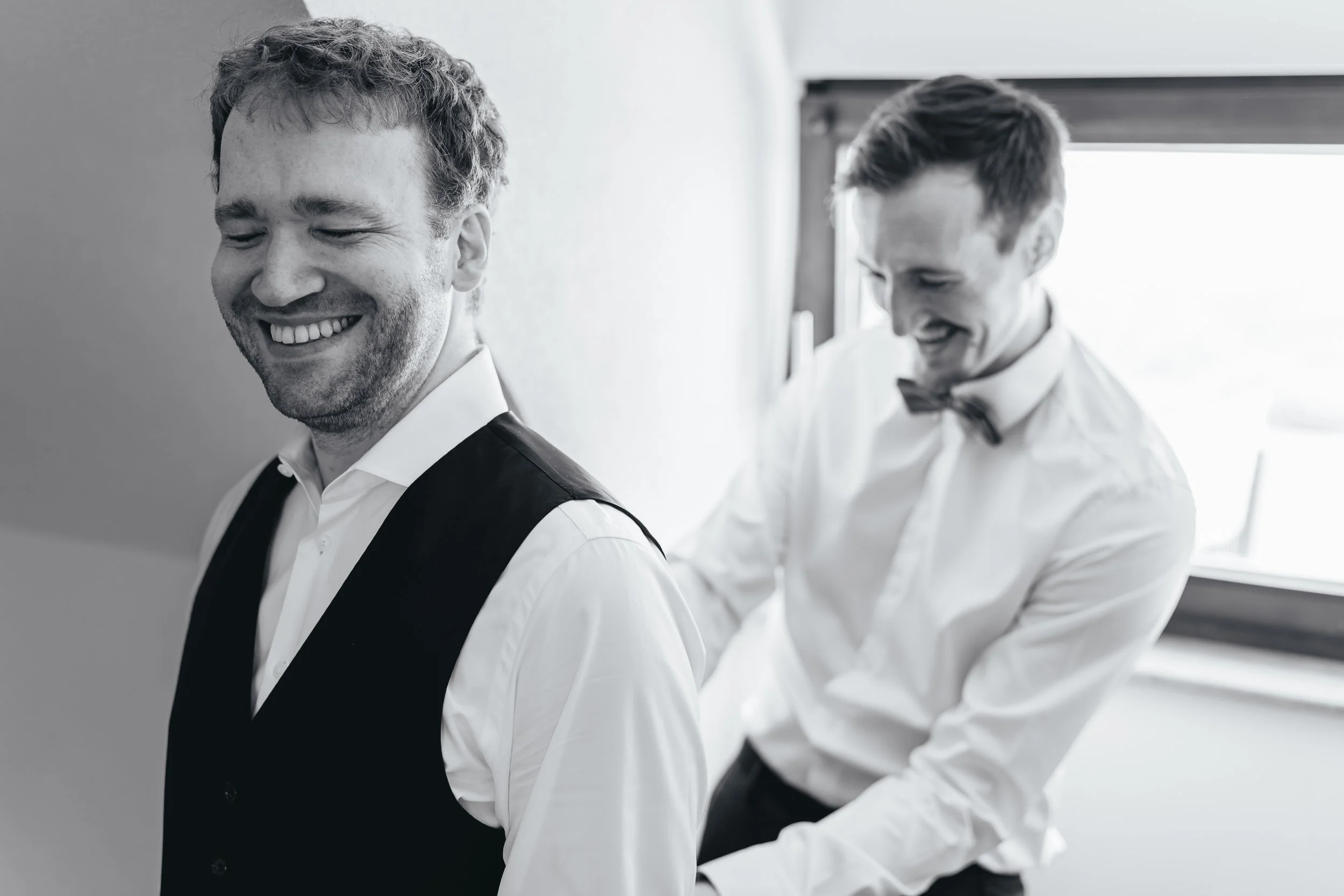 Bräutigam lacht beim anziehen, während der Trauzeuge ihm die Weste zuschnürt, schwarz weiß Foto