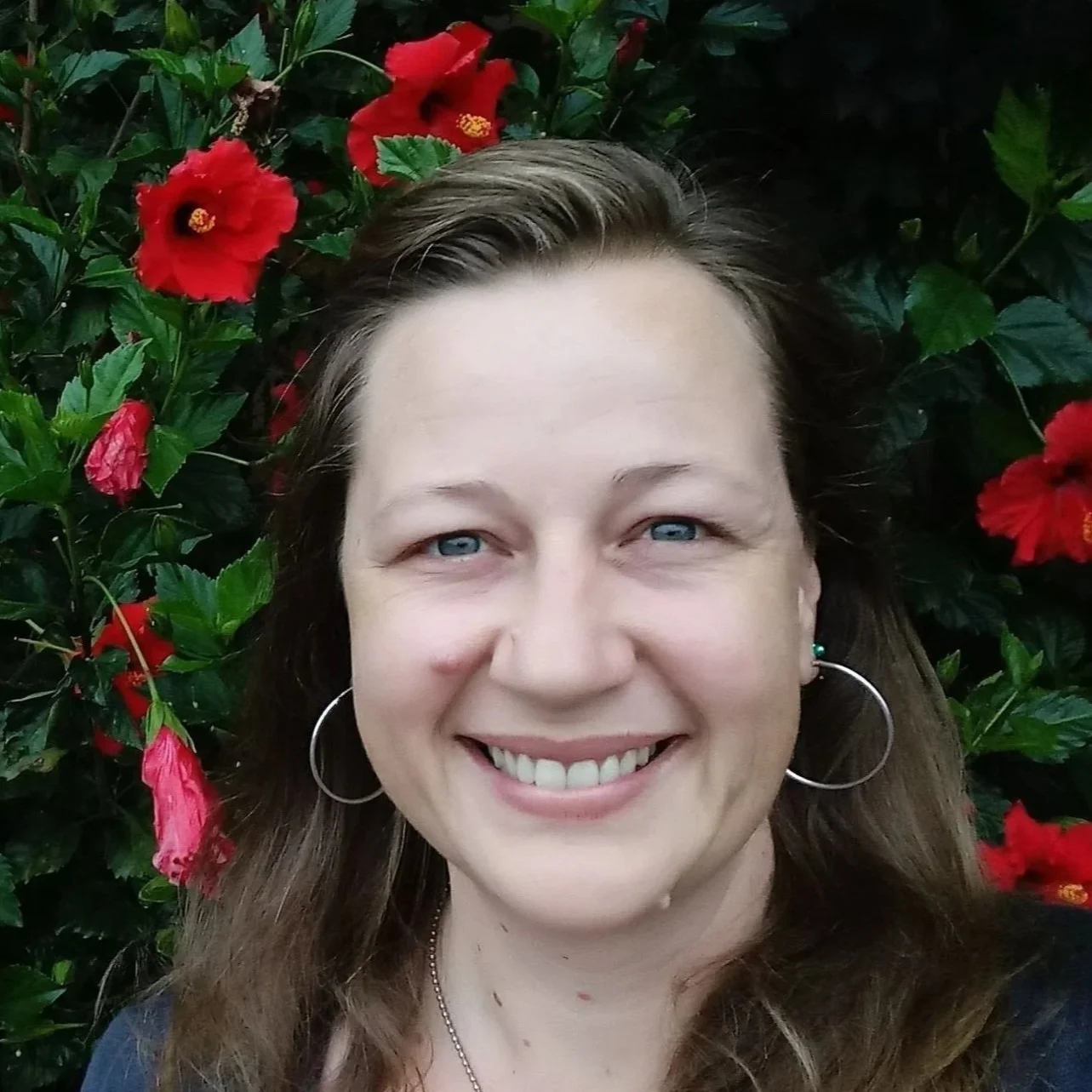 A woman smiling with blue eyes, wearing hoop earrings and a necklace, standing in front of green leaves and vibrant red hibiscus flowers.