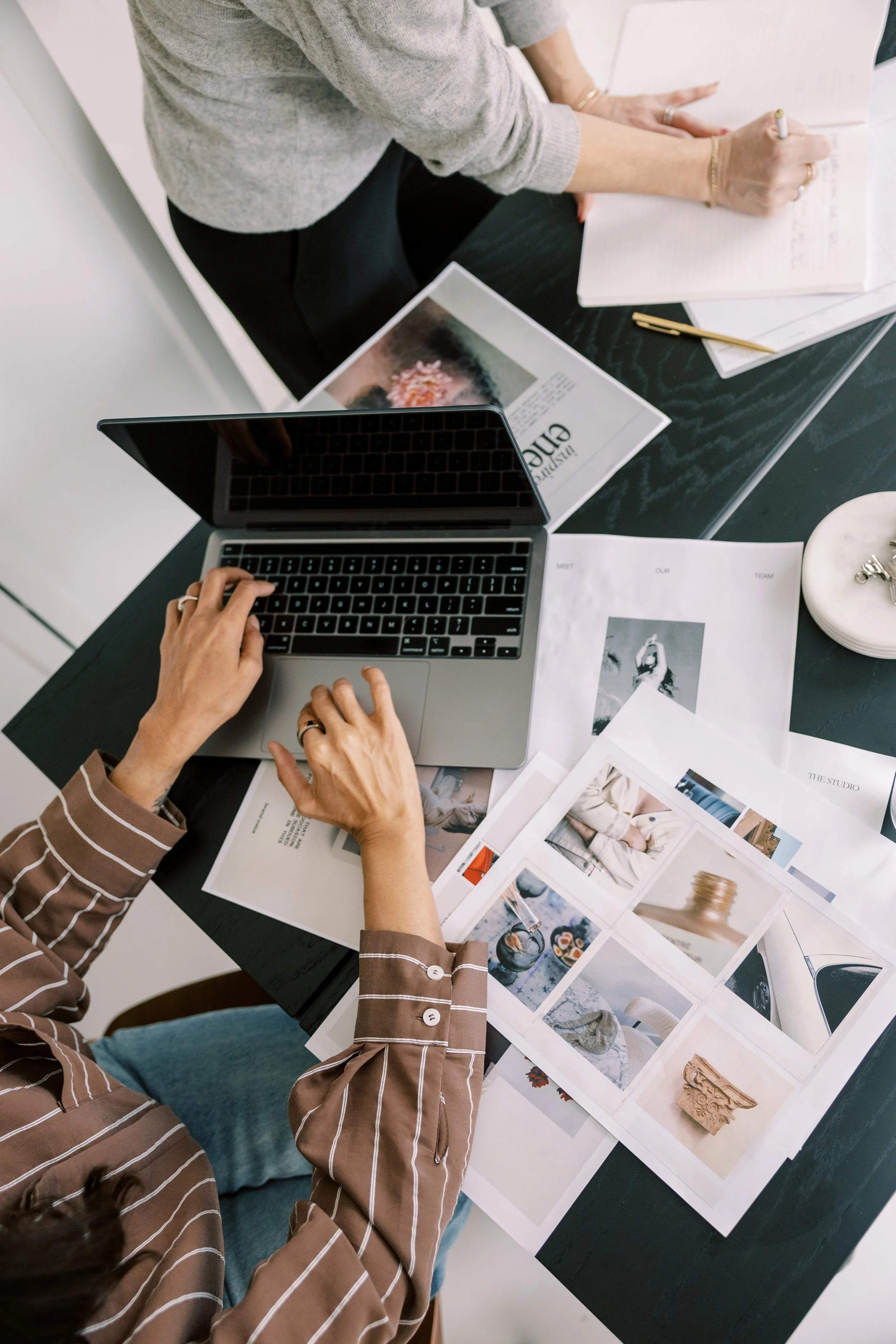 People working at a black desk with a laptop, printed photographs, magazines, and notebooks.