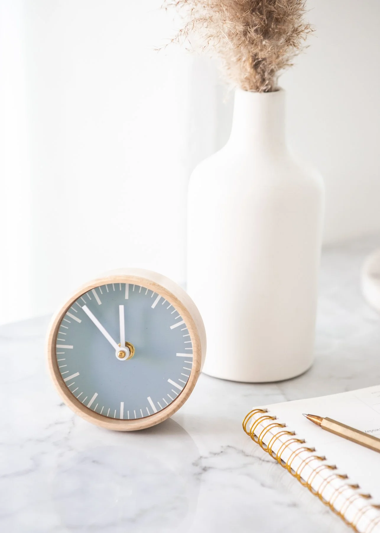 A minimalist desk setup with a gray clock, white vase with dried pampas grass, a spiral notebook, and a gold pen on a marble surface.