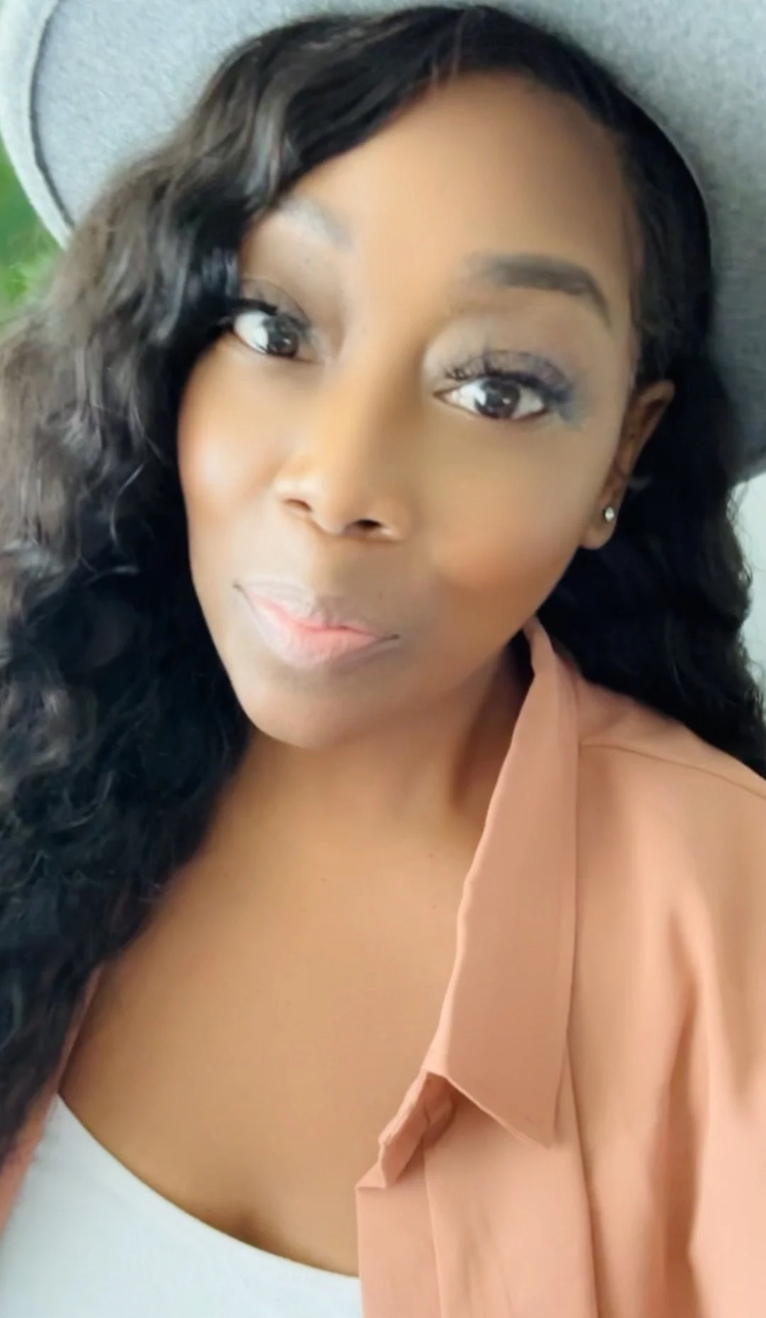 A woman wearing a gray hat and a peach-colored top, with long black hair and a neutral expression.