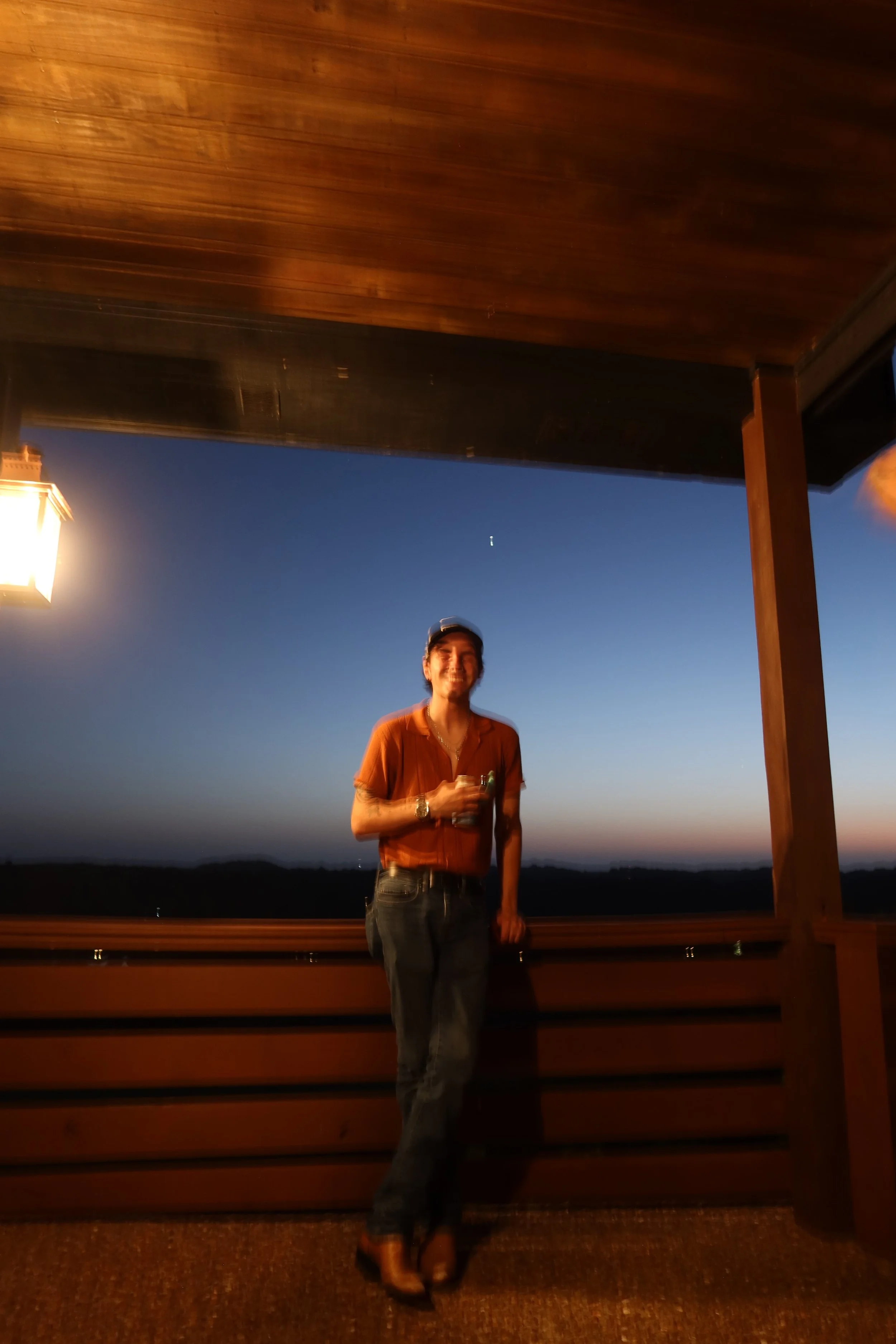 A woman standing outdoors at sunset or dusk, holding a drink in one hand, wearing a brown shirt and dark jeans, smiling, with a wooden or deck railing and a mountain or hill landscape in the background, under a ceiling with a light fixture.