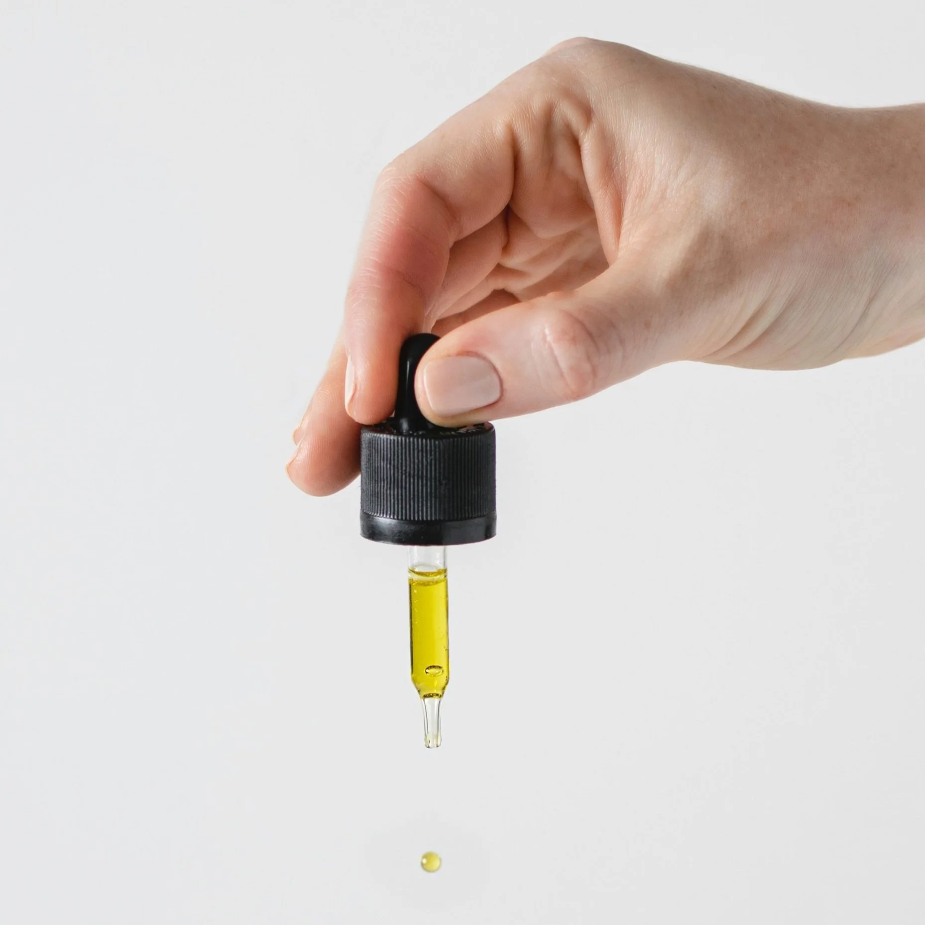 A hand holding a black dropper with yellow oil, with a droplet falling from the tip.