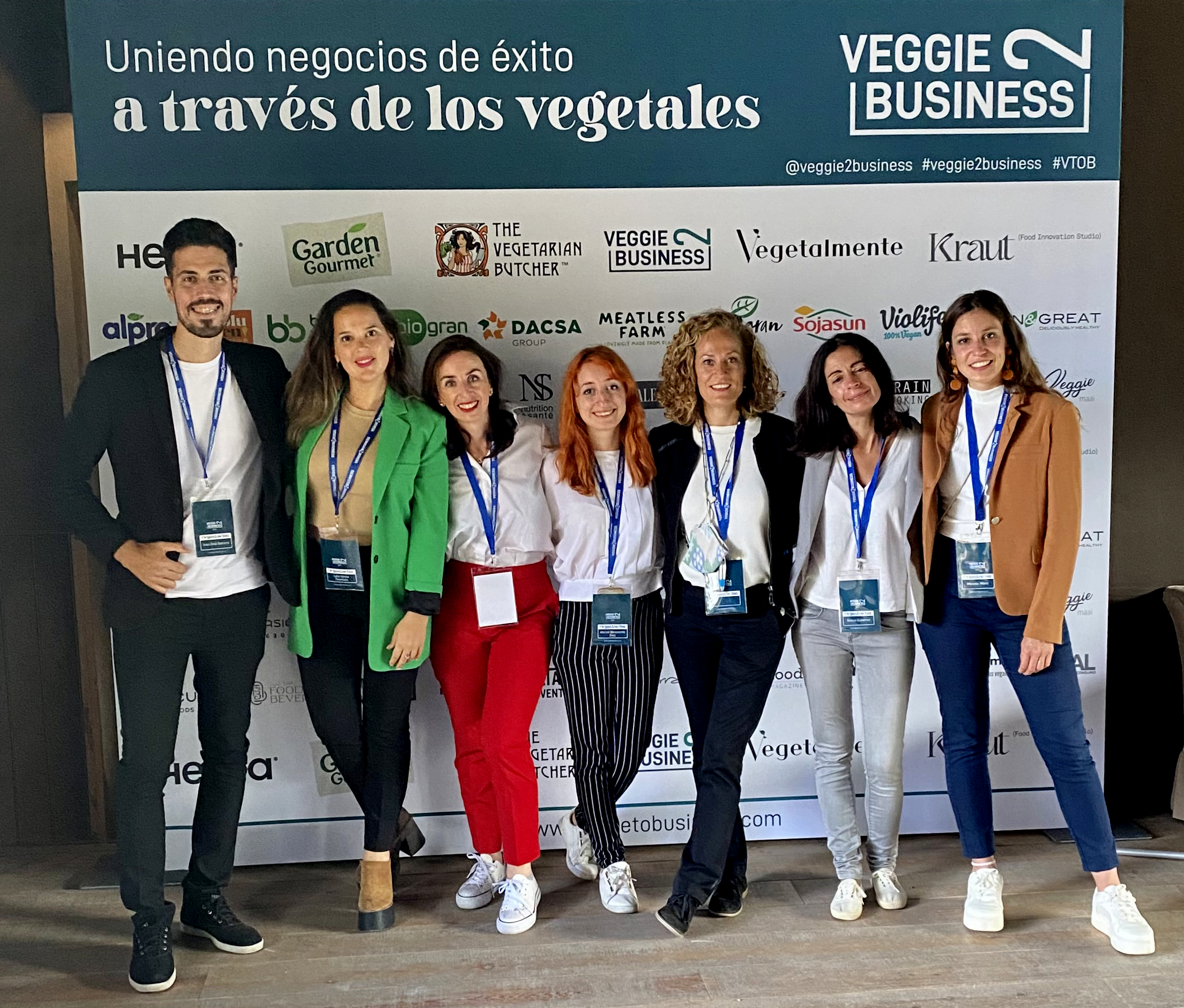 Group of seven people standing in front of a backdrop with various logos, smiling for the camera at a professional event.