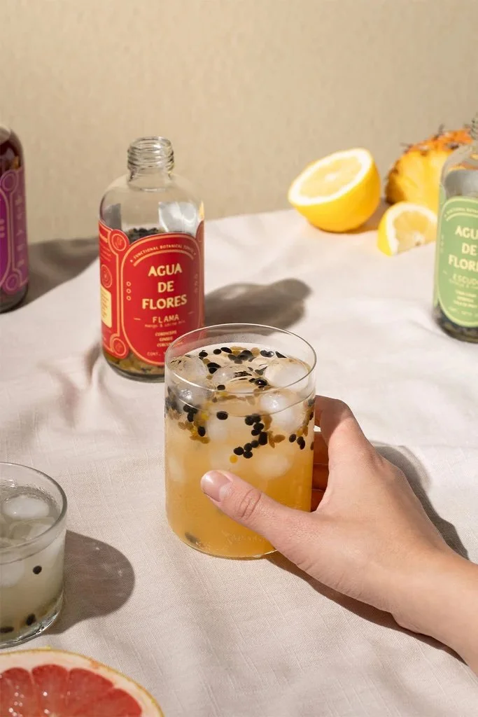 A hand holding a glass filled with ice, black seeds, and a yellow beverage, on a table with lemon slices, bottles labeled 'Agua de Flores,' and other ingredients.