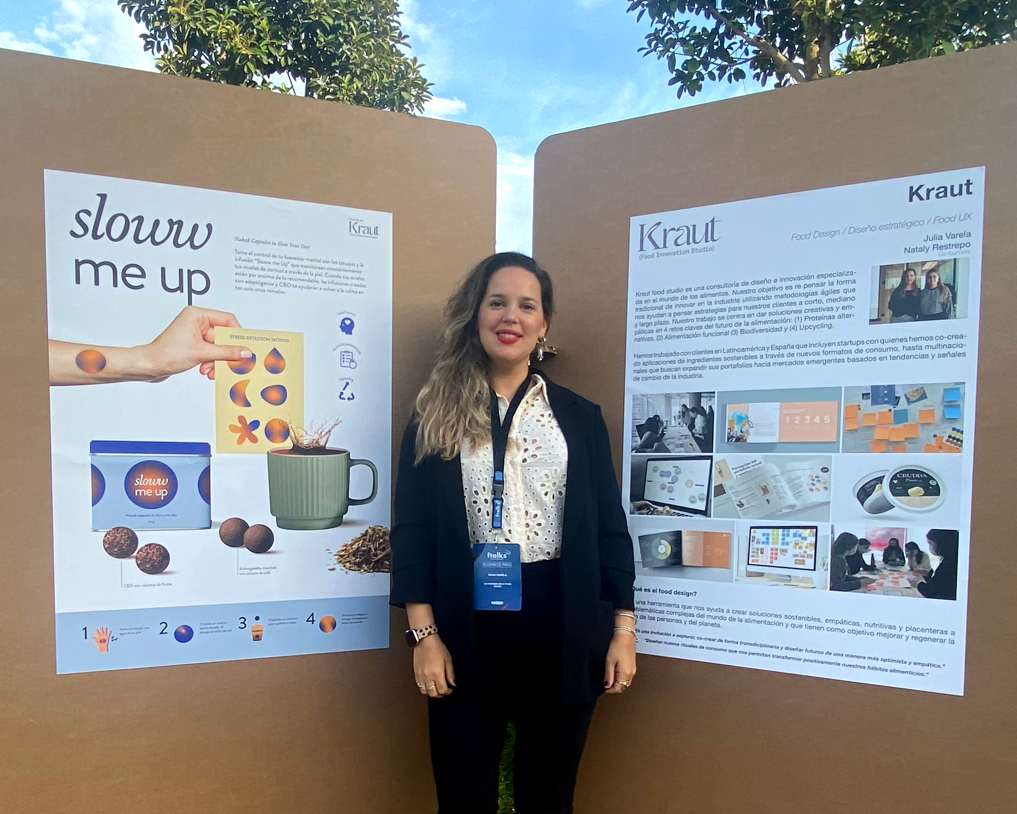 A woman standing between two display boards at an event. The left board features information about a product called 'slouw me up' with images of chocolate balls, a box, and a mug. The right board displays information about Kraut food design, including images of creative food packaging and design projects.