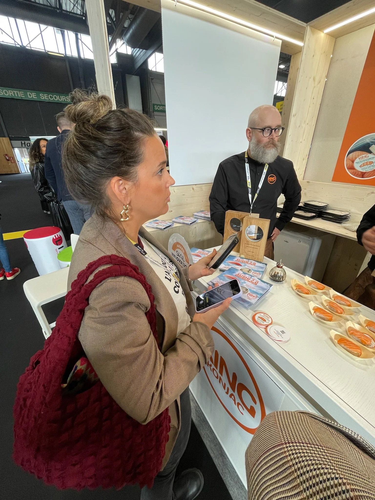 Woman with a brown blazer and red textured vest, holding two smartphones, standing at a booth with sushi samples, at an event or exhibition.