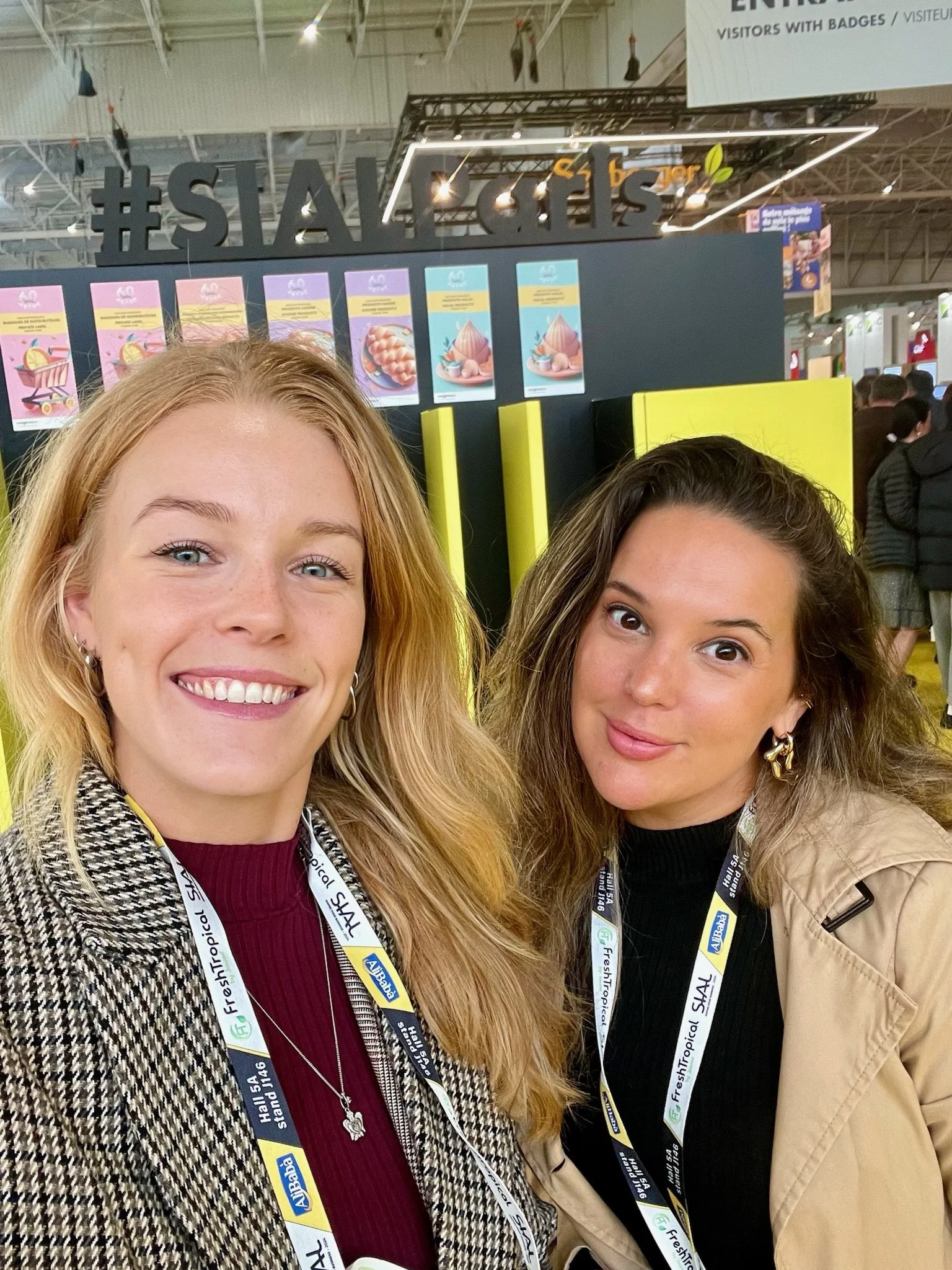 Two women smiling for a selfie at a trade show or exhibition, with lanyards around their necks and a display with colorful posters and large hashtag sign in the background.