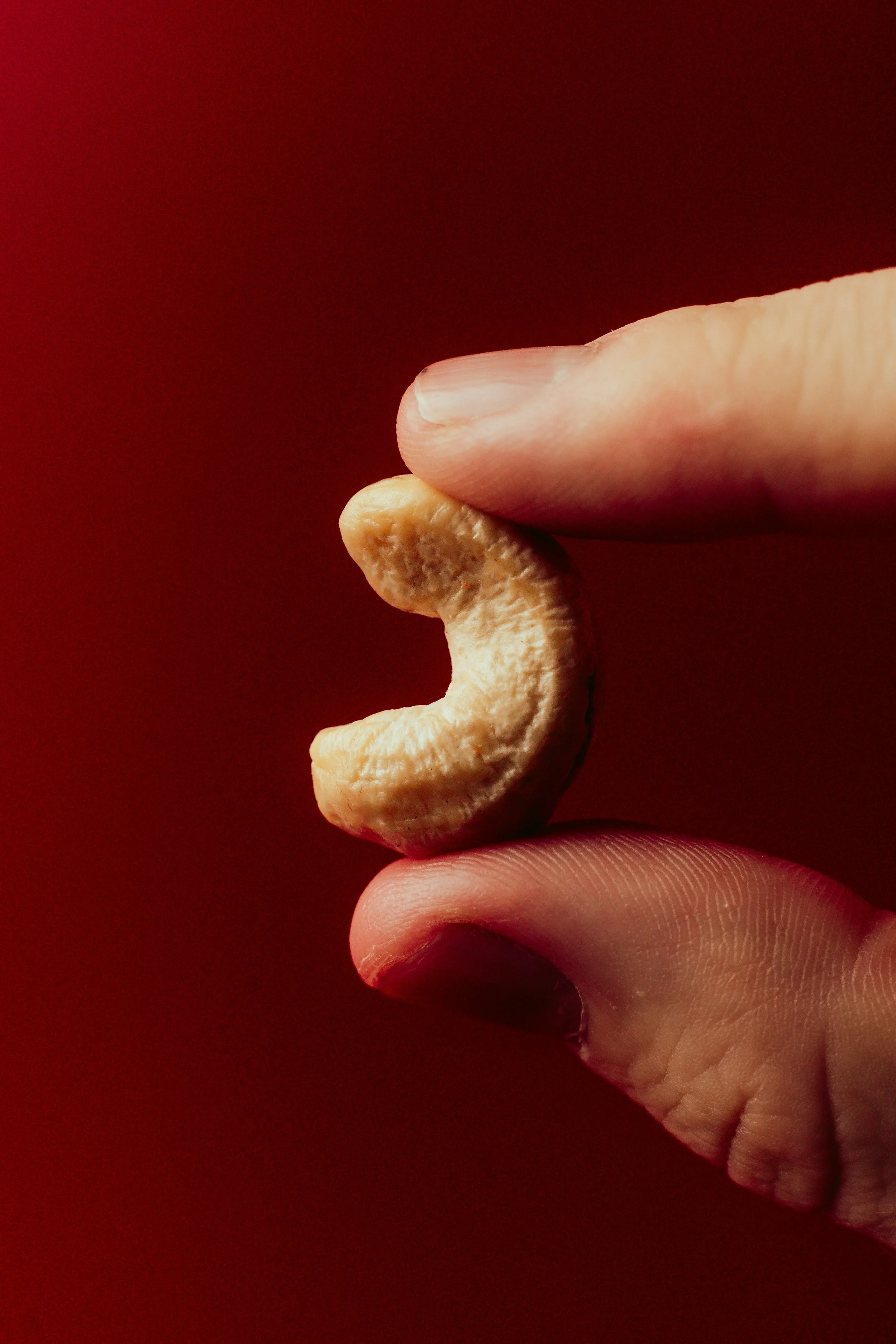 Close-up of a hand holding a single cashew nut against a dark red background.