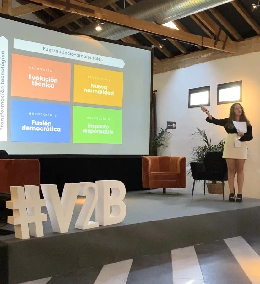 A woman giving a presentation in a room with a large screen displaying four scenarios about socio-environmental forces. The scenarios are titled 'Evolución técnica,' 'Nueva normalidad,' 'Fusión democrática,' and 'Impacto responsable,' each with different colors. There are large white #V2B letters on the stage in front of her, and the room has wooden beams, plants, and two small windows.