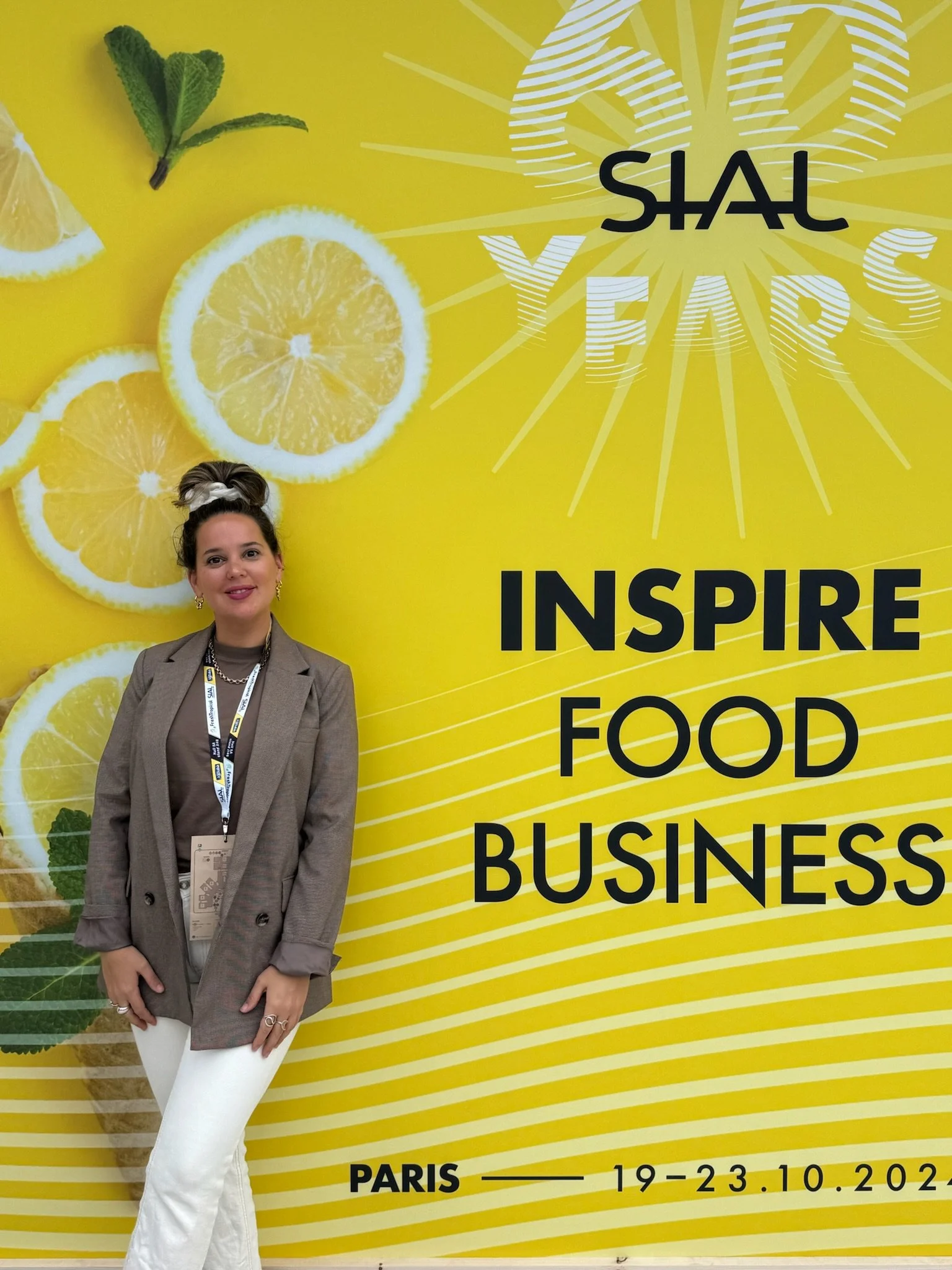 A woman standing in front of a bright yellow wall with lemon slices and a logo that says 'SHALE Y EARS.' The wall has the phrases 'INSPIRE FOOD BUSINESS' and 'PARIS 19-23.10.2022' written on it. The woman is wearing a beige blazer, a brown top, white pants, and has a lanyard around her neck.