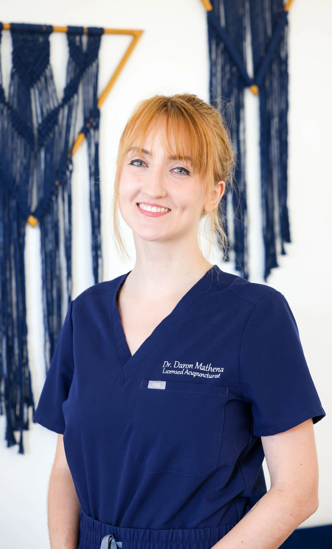 A smiling woman with red hair wearing navy scrubs with the text 'Dr. Daron Mathena, Licensed Acupuncturist.' She is standing in front of a white wall decorated with navy blue woven wall hangings.