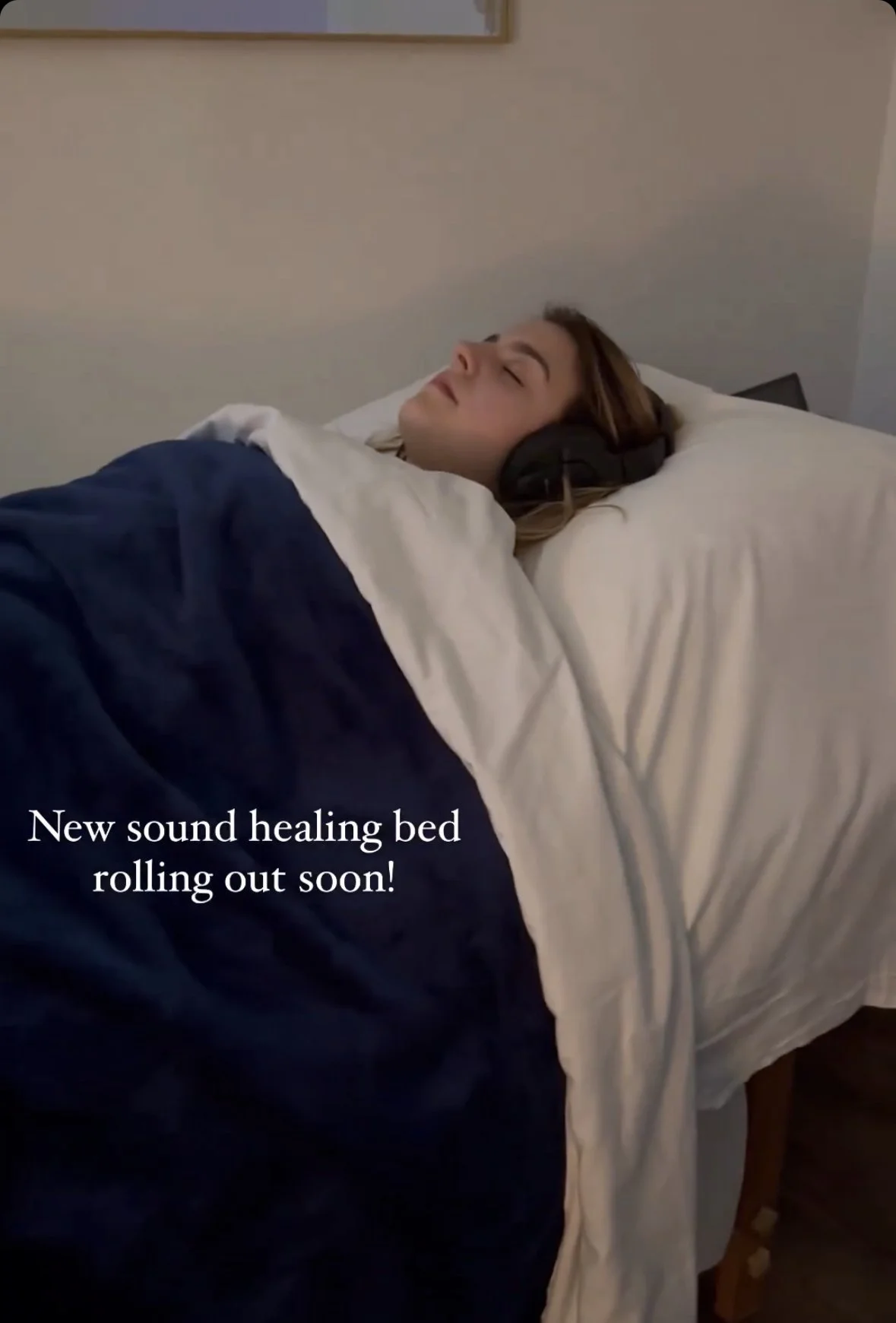 A woman with headphones sleeping on a bed with a white pillow and dark blue blanket, with text overlay saying 'New sound healing bed rolling out soon!'