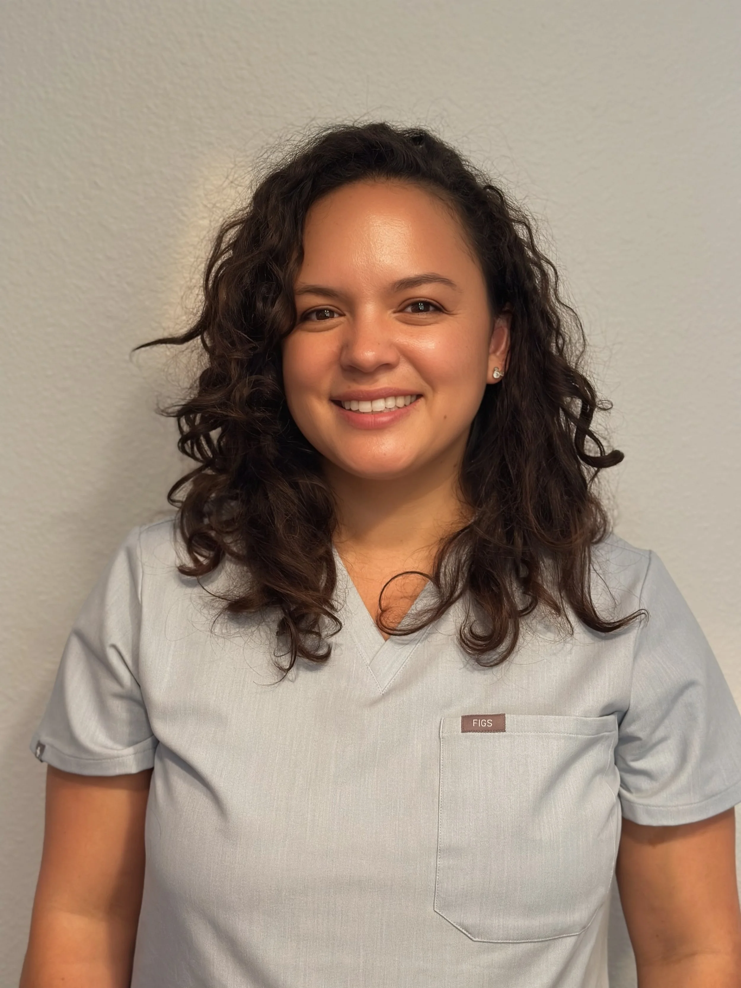A woman with curly dark hair wearing light gray medical scrubs standing against a plain wall, smiling.