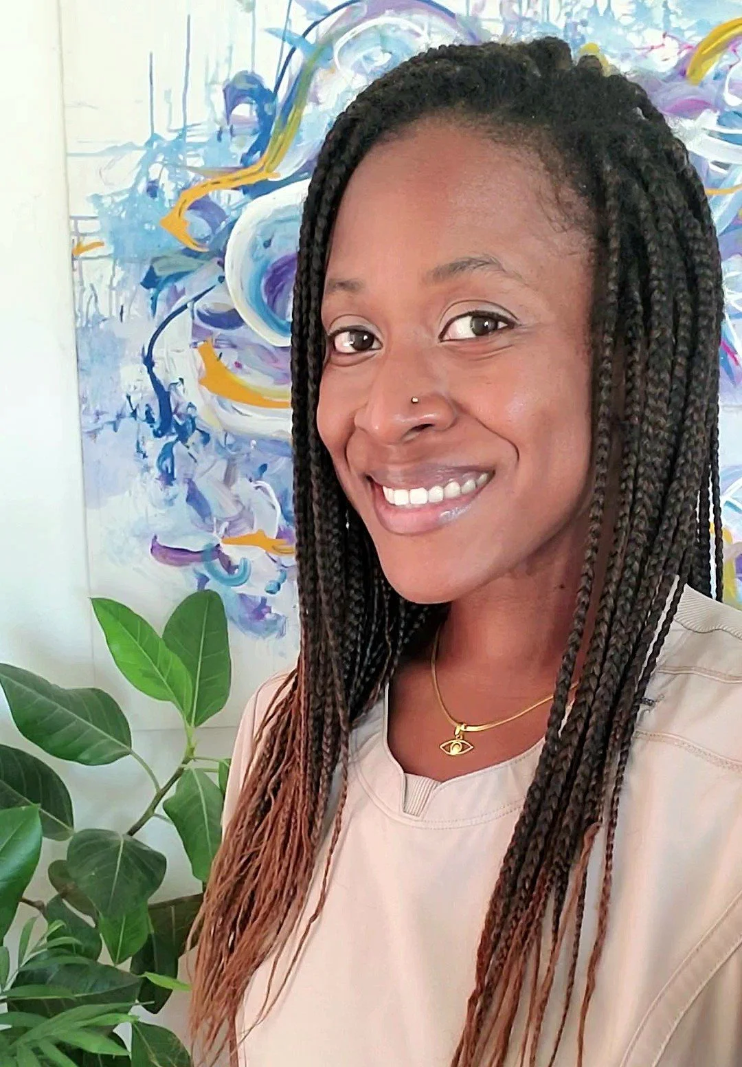 A woman with braided hair smiling in front of an abstract painting with purple, blue, yellow, and white swirls, and green leaves in the foreground.