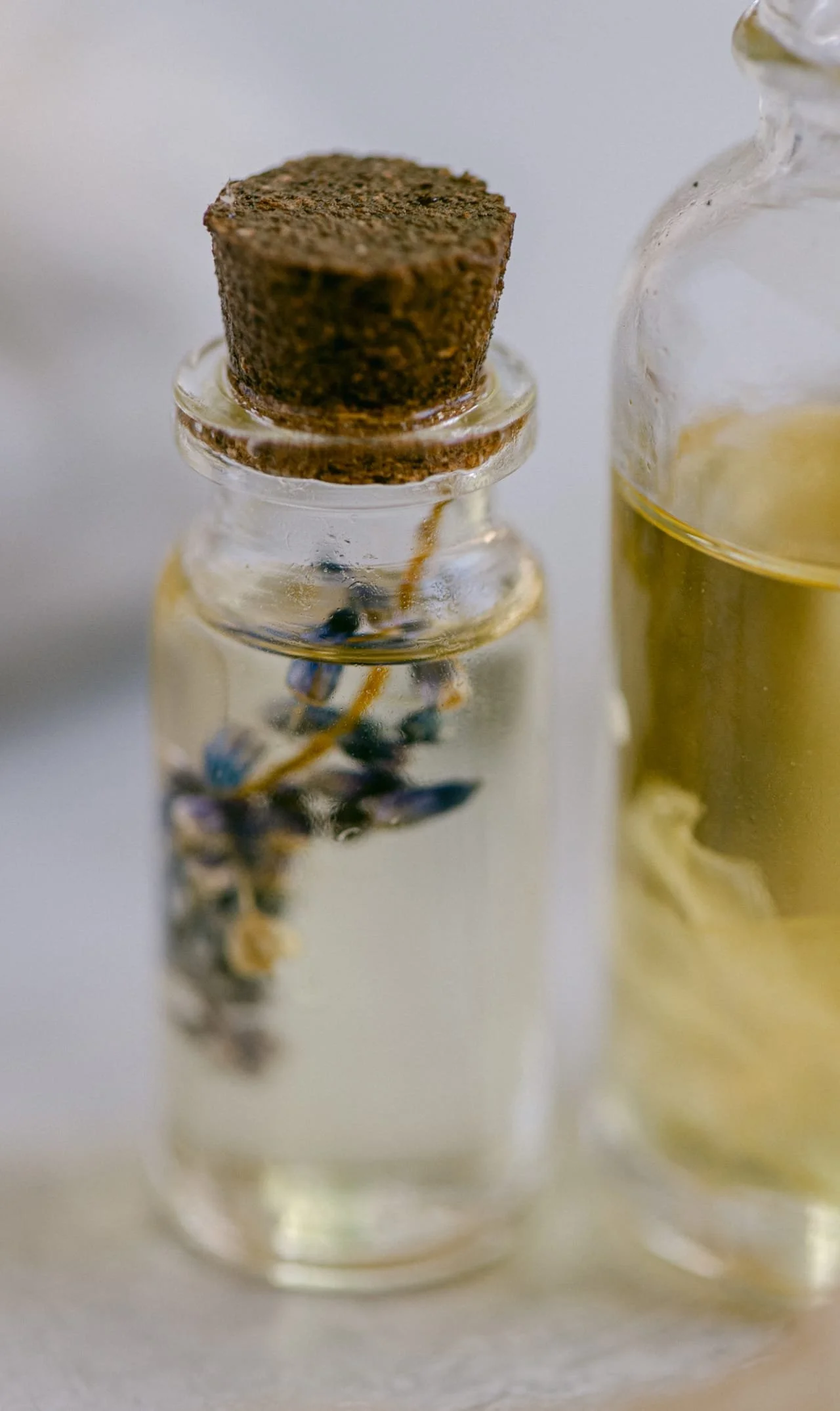 A small glass bottle with a cork stopper, containing dried lavender flowers and a yellowish liquid, possibly essential oil, next to another similar bottle.