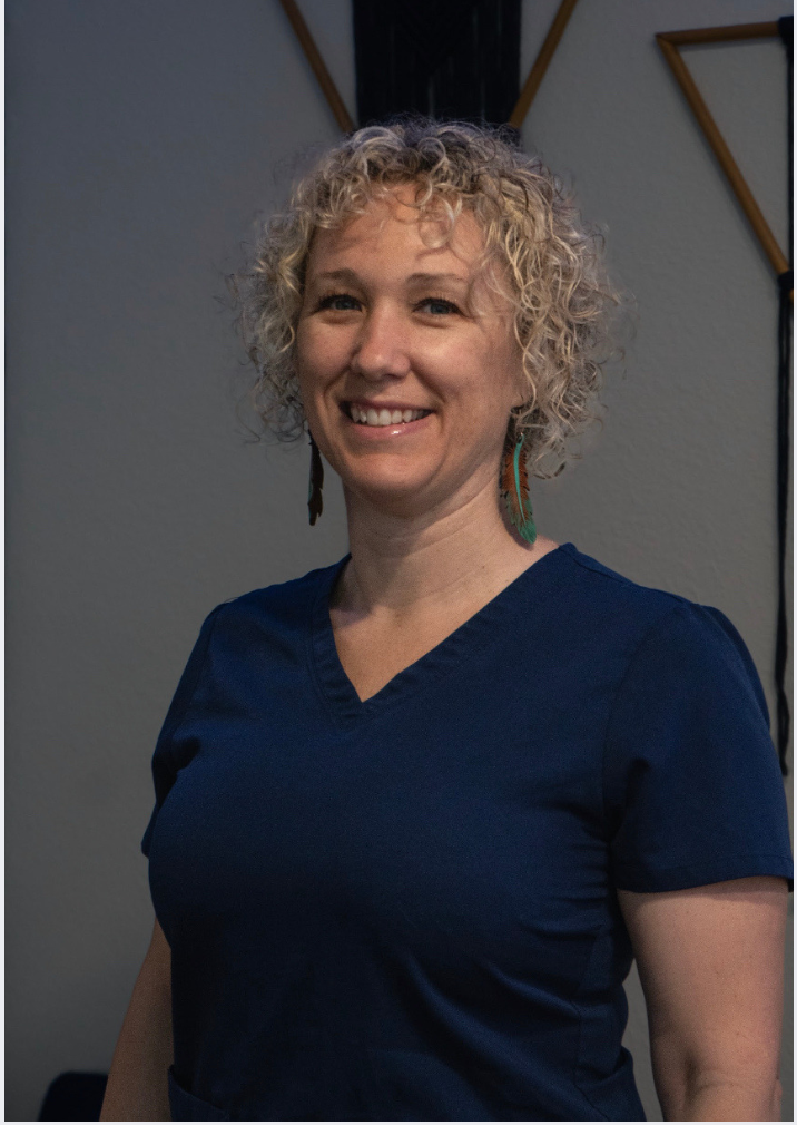 A woman with curly blonde hair wearing a dark blue shirt, smiling, standing in front of a gray wall with some geometric wall decorations.