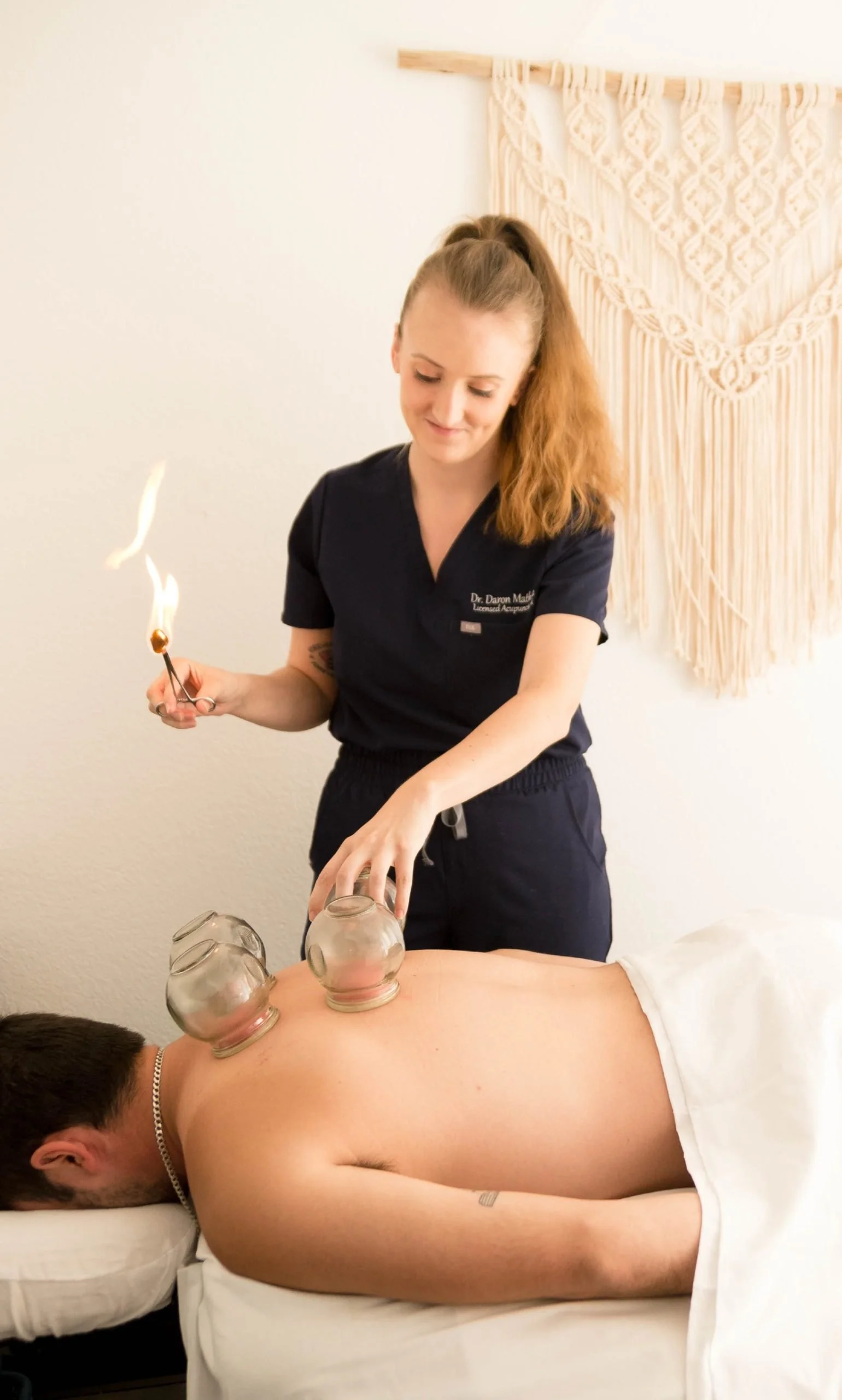 A woman performing a glass cupping therapy on a man lying face down on a massage table, with cups on his back and a therapist holding a flaming matchstick.
