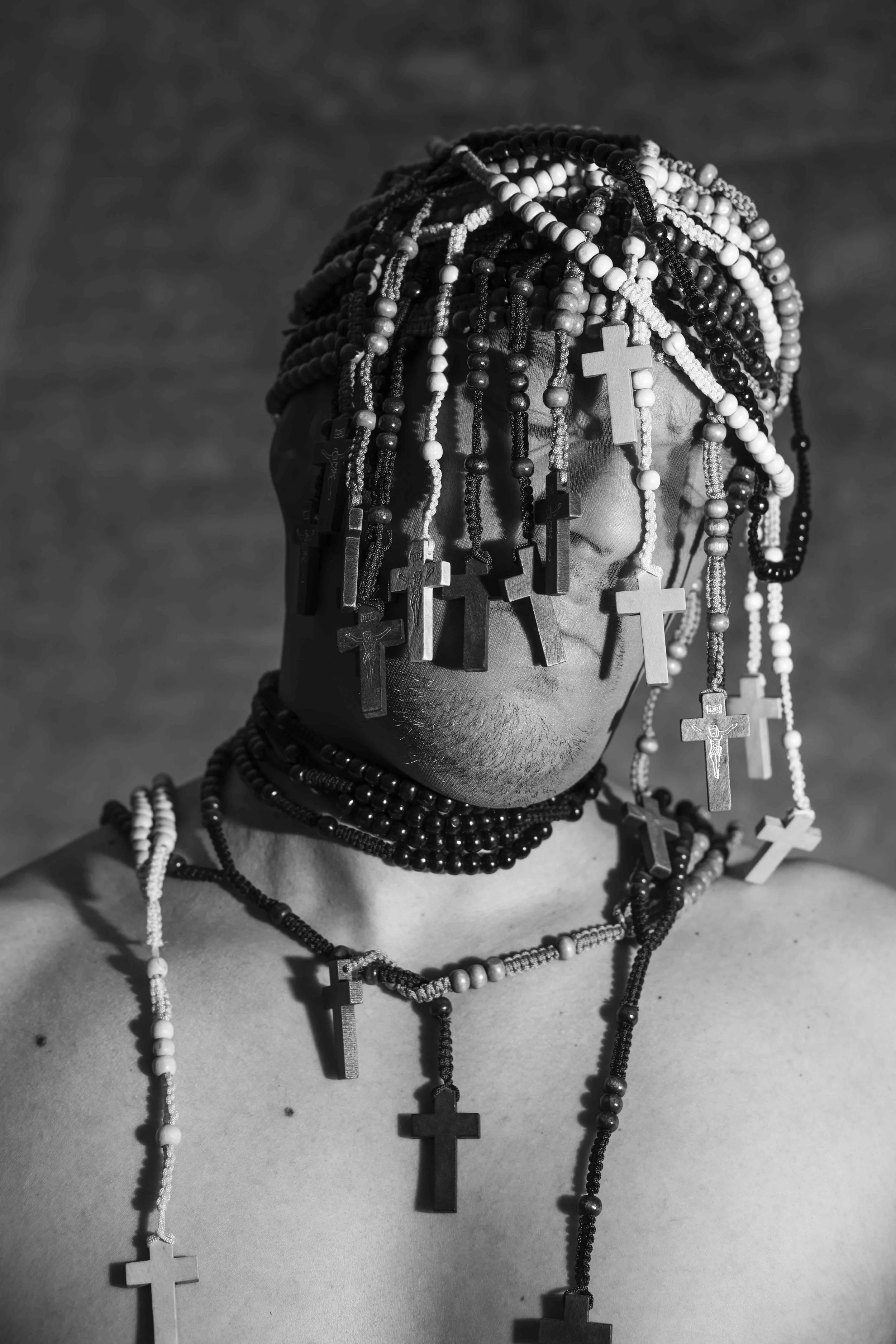 Black and white photo of a person adorned with numerous beaded necklaces and rosaries with crosses, covering their head and face.