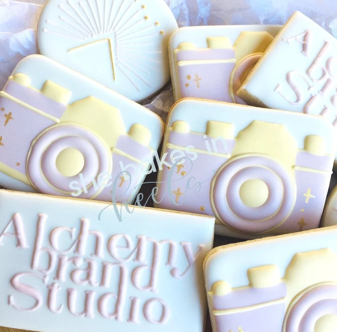 Decorative cookies featuring designs of cameras and the text "Alchemy Brand Studio" with pastel colors.