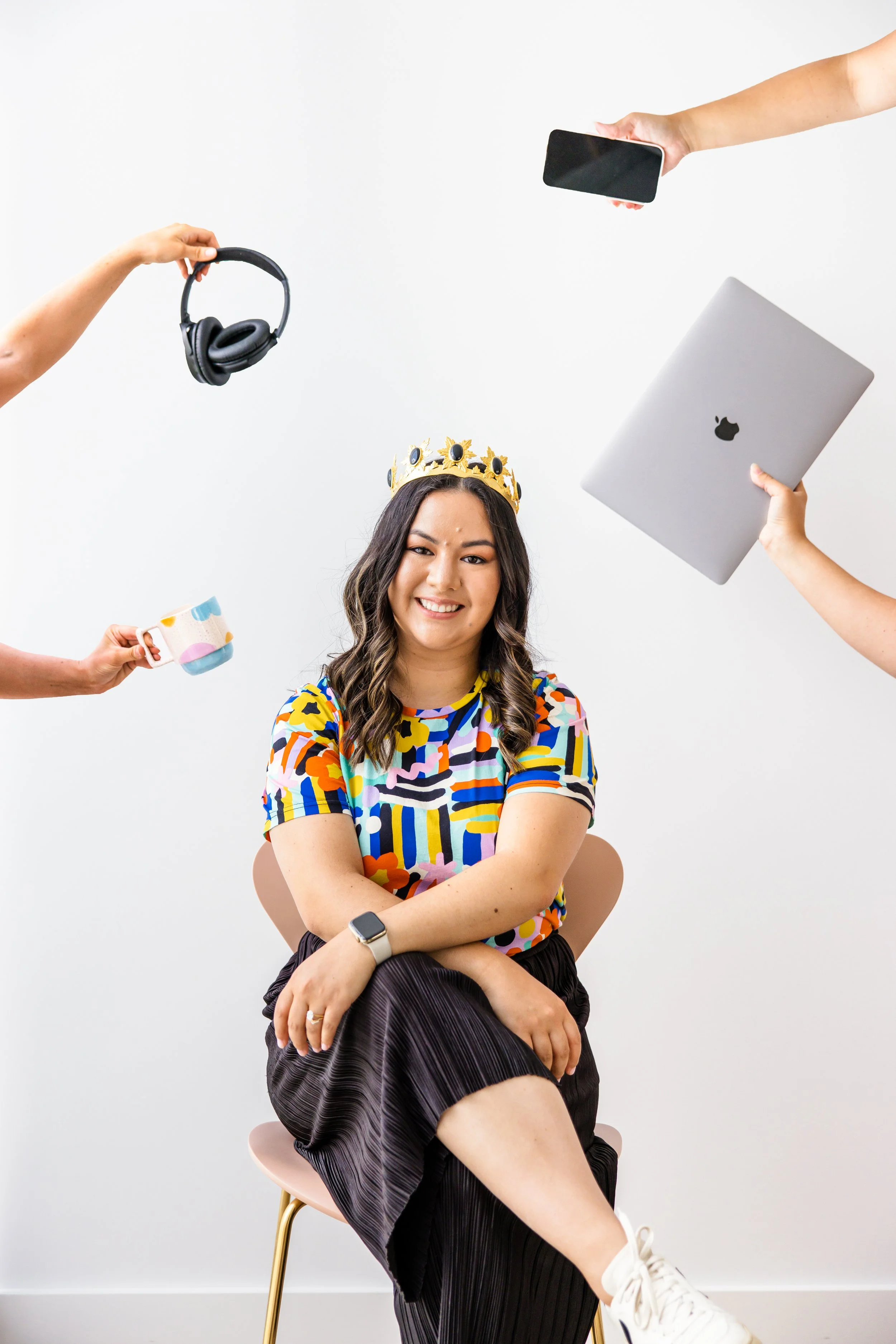 Woman wearing a colorful shirt and a crown, sitting on a chair, with four arms holding headphones, a mug, a phone, and a laptop around her.
