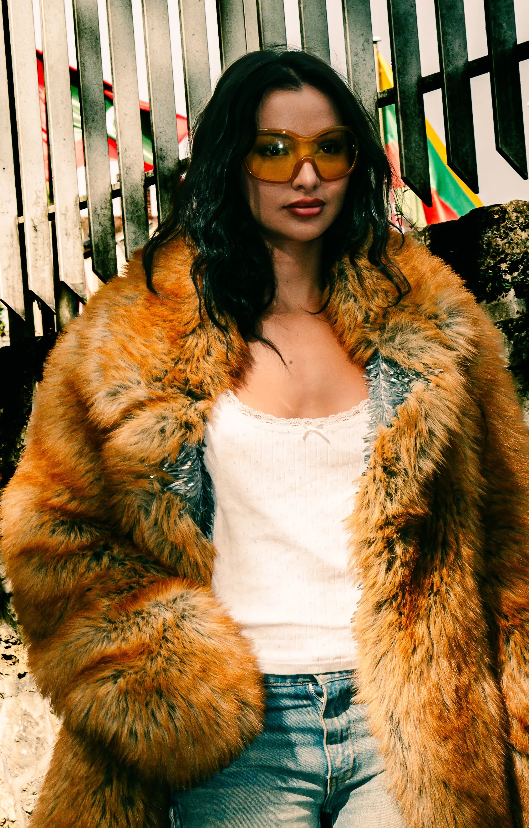 Une femme porte un manteau en fourrure, des lunettes de soleil jaunes, un t-shirt blanc et un jean, devant une structure en métal.
