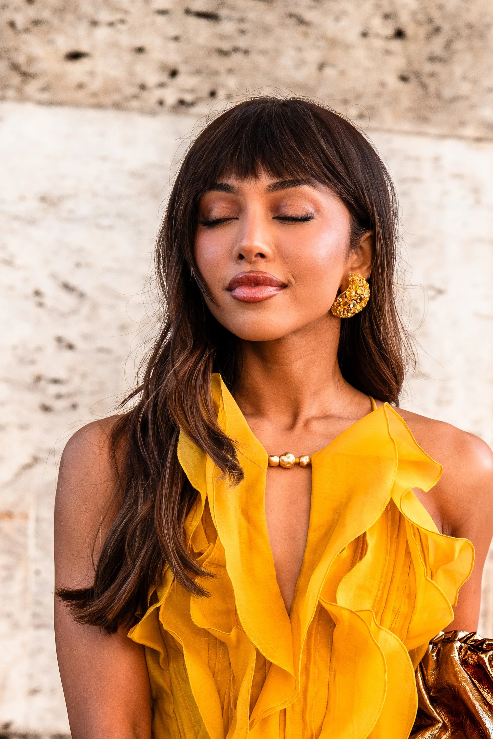 Une femme portant une robe jaune avec beaucoup de détails plissés et ruffles, portant de grosses boucles d'oreilles dorées et un collier de perles. Son visage est détendu avec les yeux fermés, dans un contexte intérieur avec un mur en pierre en arriè