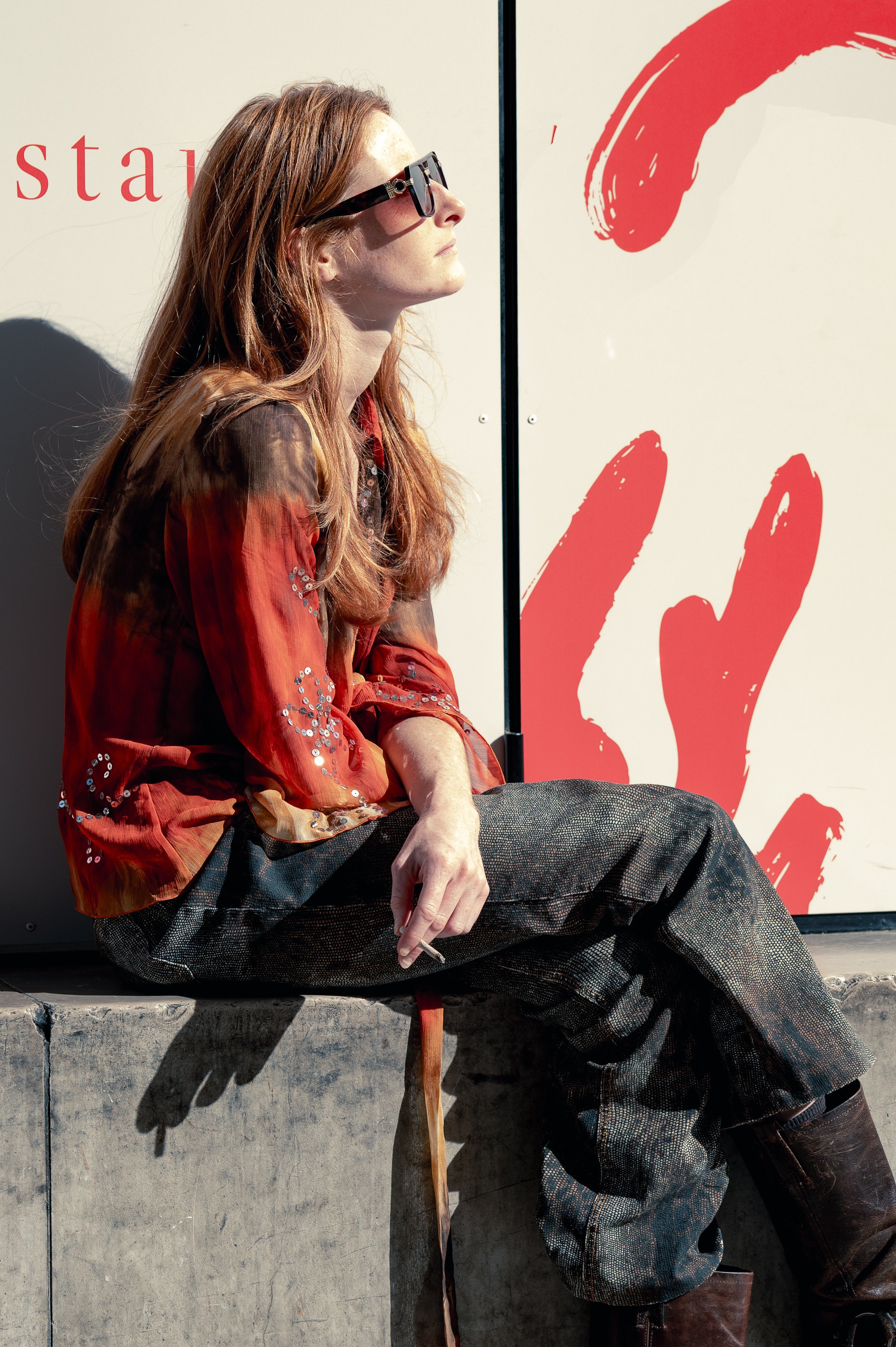 Une femme assise sur un rebord en béton, portant des lunettes de soleil, une chemise rouge avec des détails brillants, des pantalons larges à carreaux, et des bottes en cuir, avec un fond blanc et rouge artistiquement peint.
