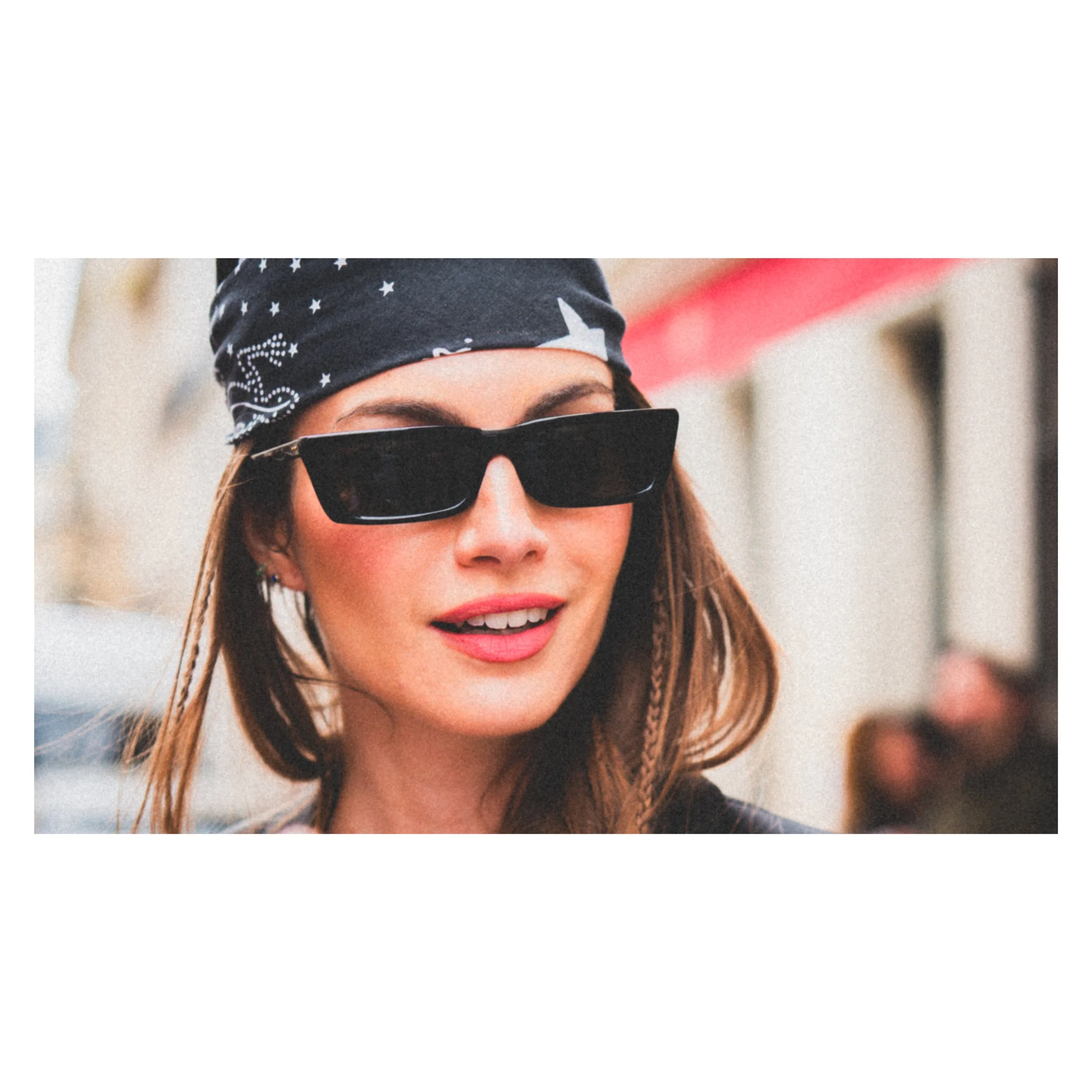 Une femme souriante portant des lunettes de soleil noires, un bandana noir avec des motifs de stars, et des cheveux longs et bruns.