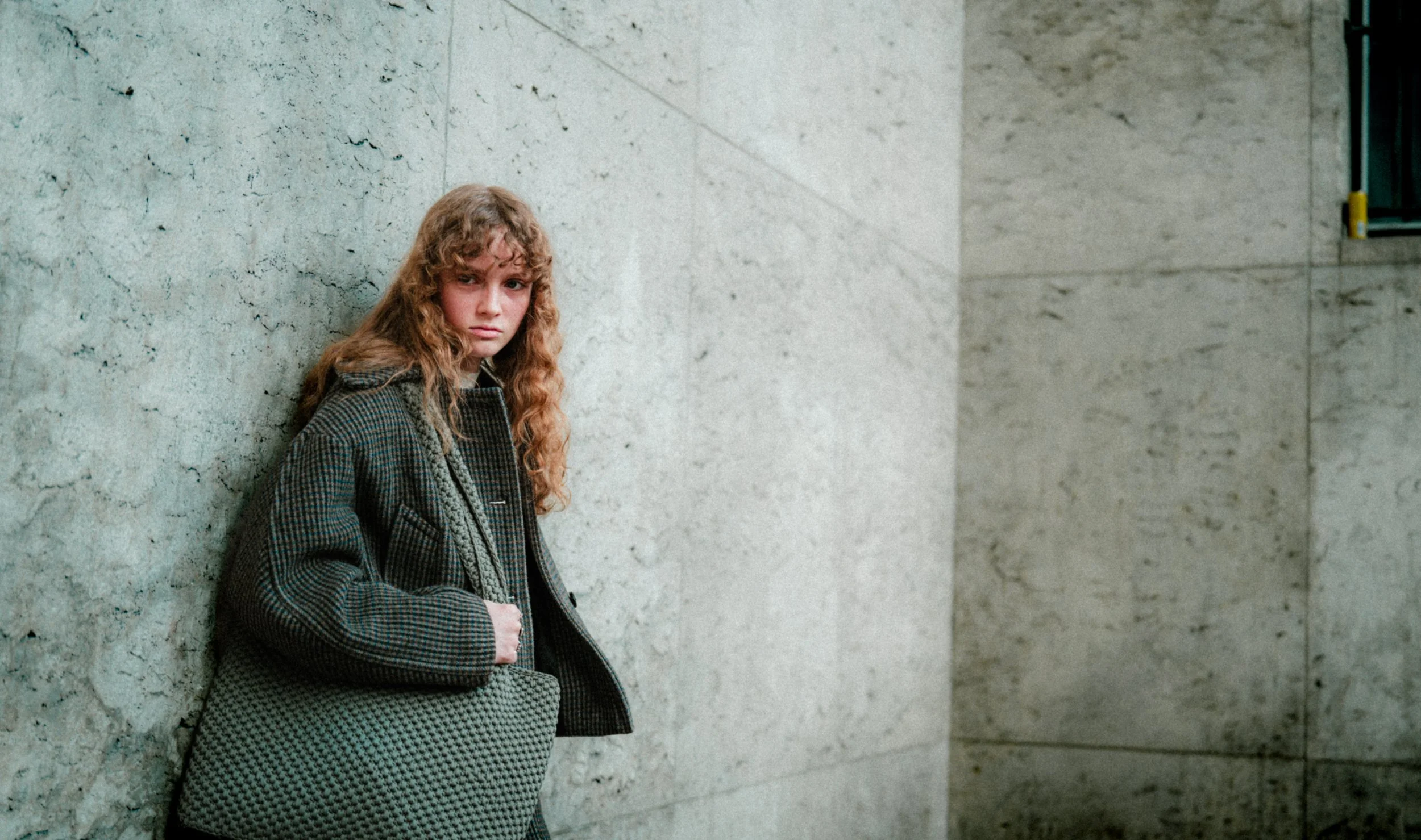Jeune femme aux cheveux frisés, vêtue d'un manteau à carreaux, appuyée contre un mur en pierre dans un environnement urbain.