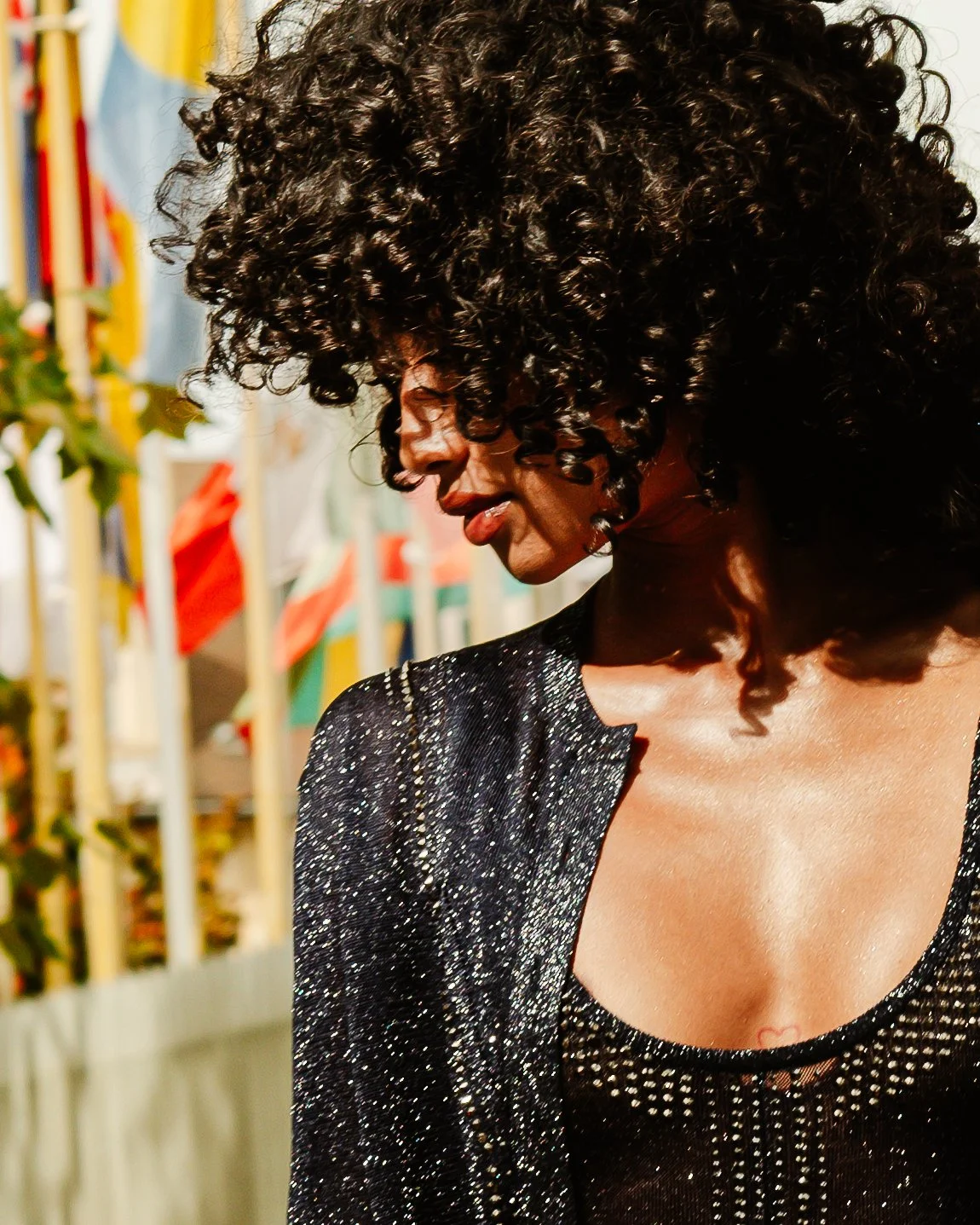 Une femme avec des cheveux bouclés noirs, portant un vêtement noir brillant avec des perles, est de profil et semble être à une fête ou un événement extérieur.