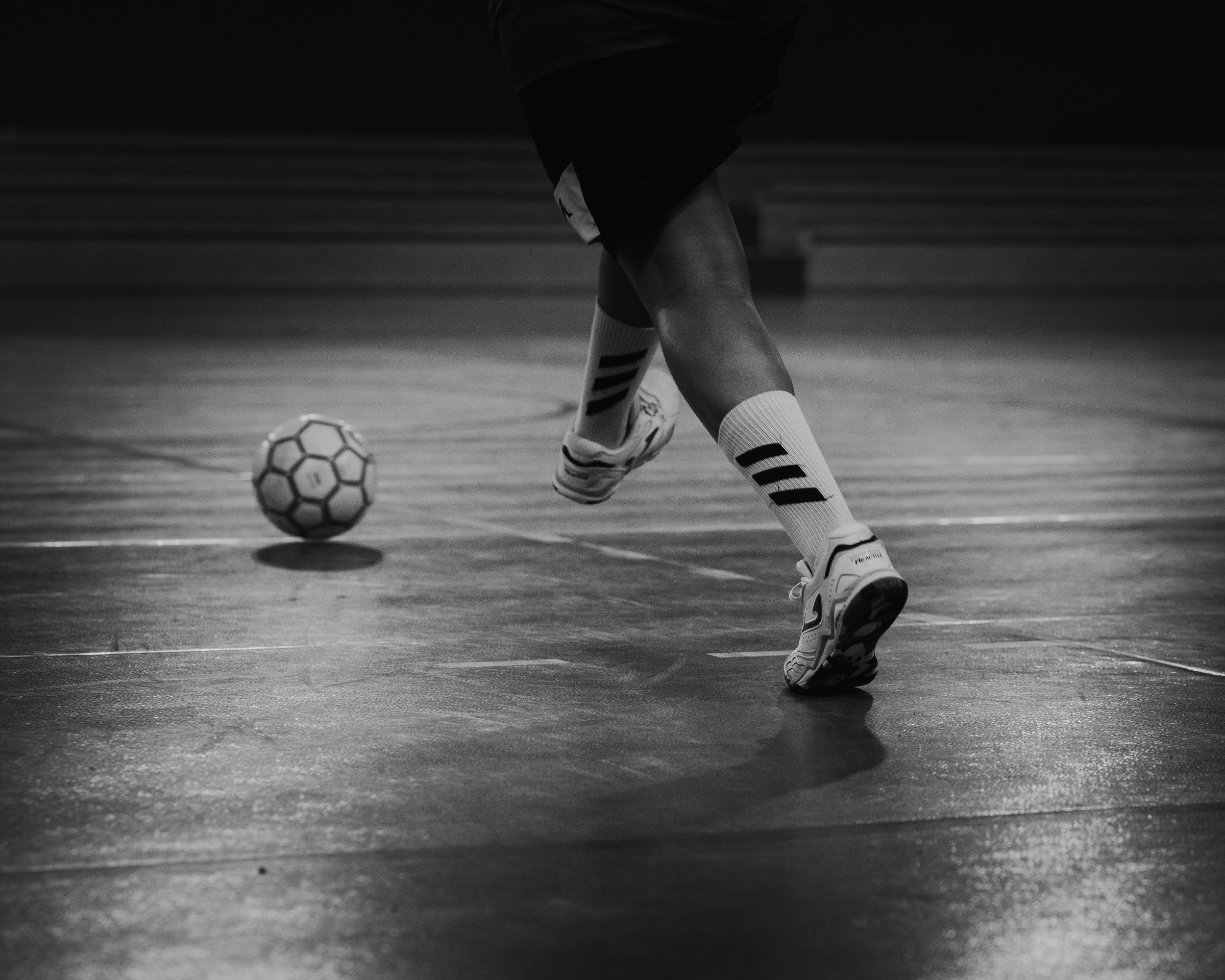 Un joueur de soccer en mouvement sur un terrain intérieur, avec un ballon à proximité, en noir et blanc.
