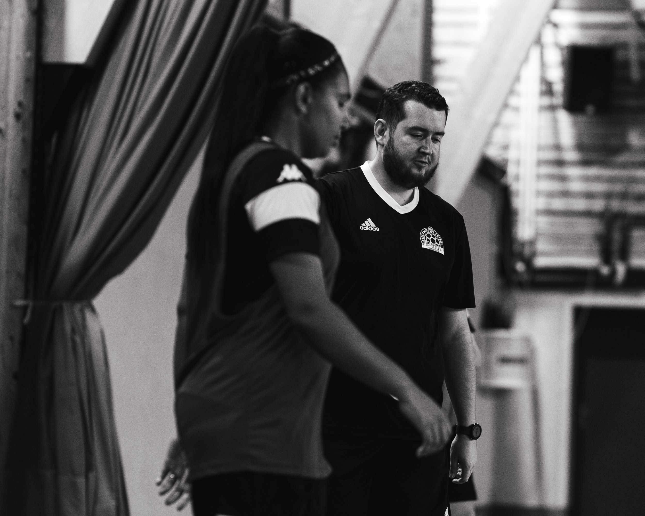 Deux personnes en tenue sportive discutent dans une salle, l'une en avant-plan et l'autre en arrière-plan.