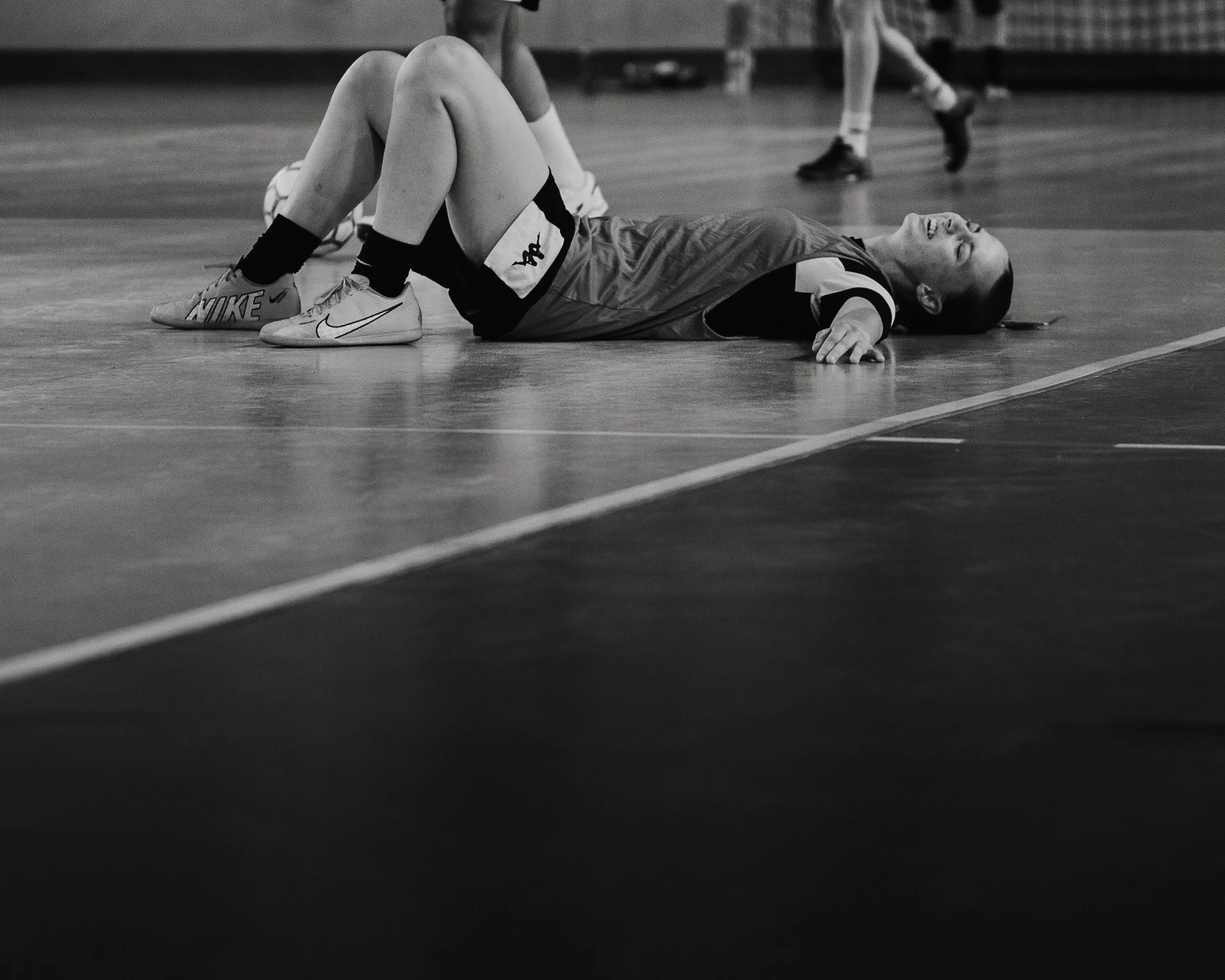 Une fille allongée sur un sol de gym ou de sport, visiblement blessée ou essoufflée, portant une tenue de sport et des chaussures de sport. Elle est dans un gymnase ou un terrain de sport, avec d'autres personnes en arrière-plan.