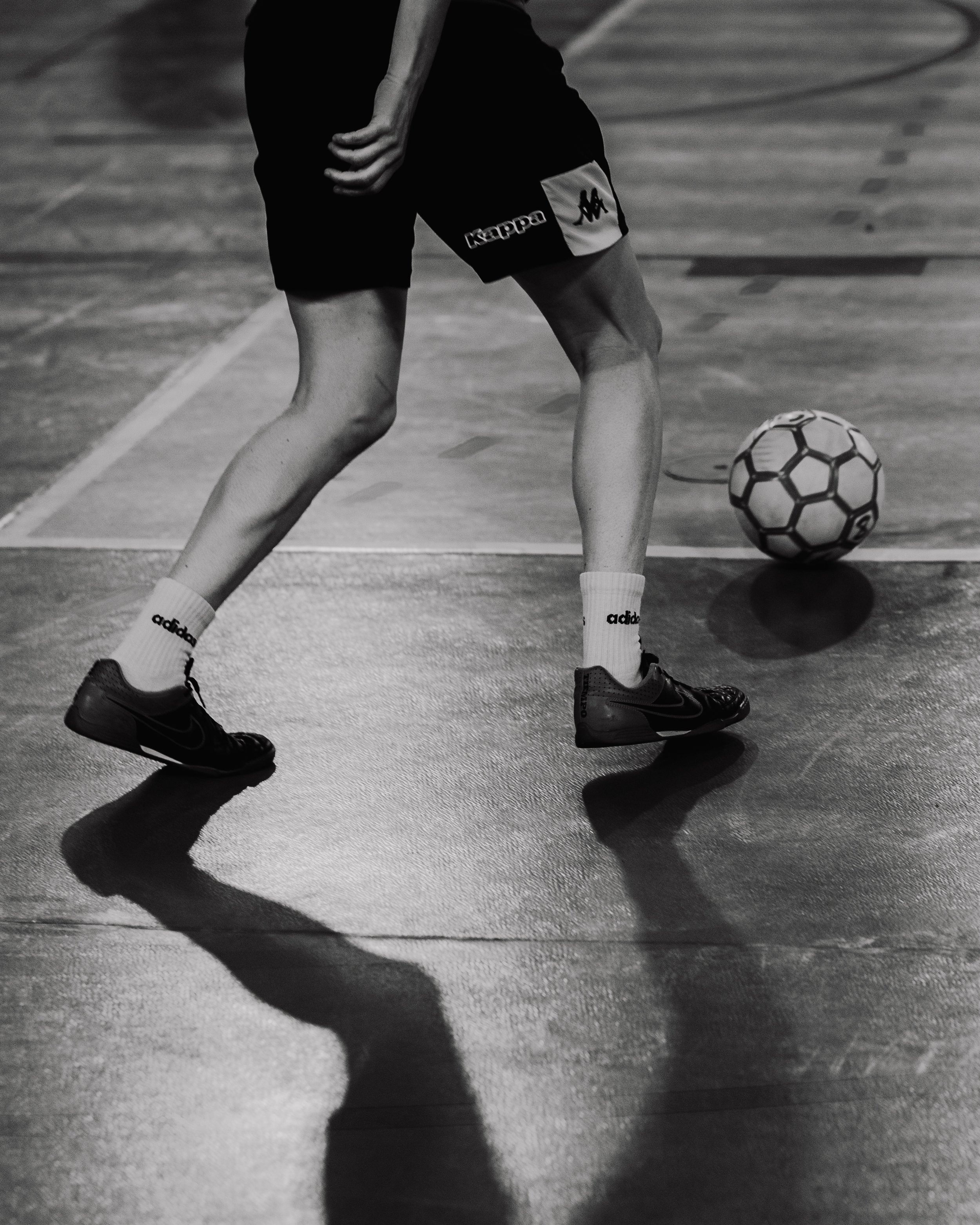 Un enfant joue au football en intérieur, portant des baskets Nike, des chaussettes Adidas, et un short avec un logo Kappa, avec un ballon de football devant lui.