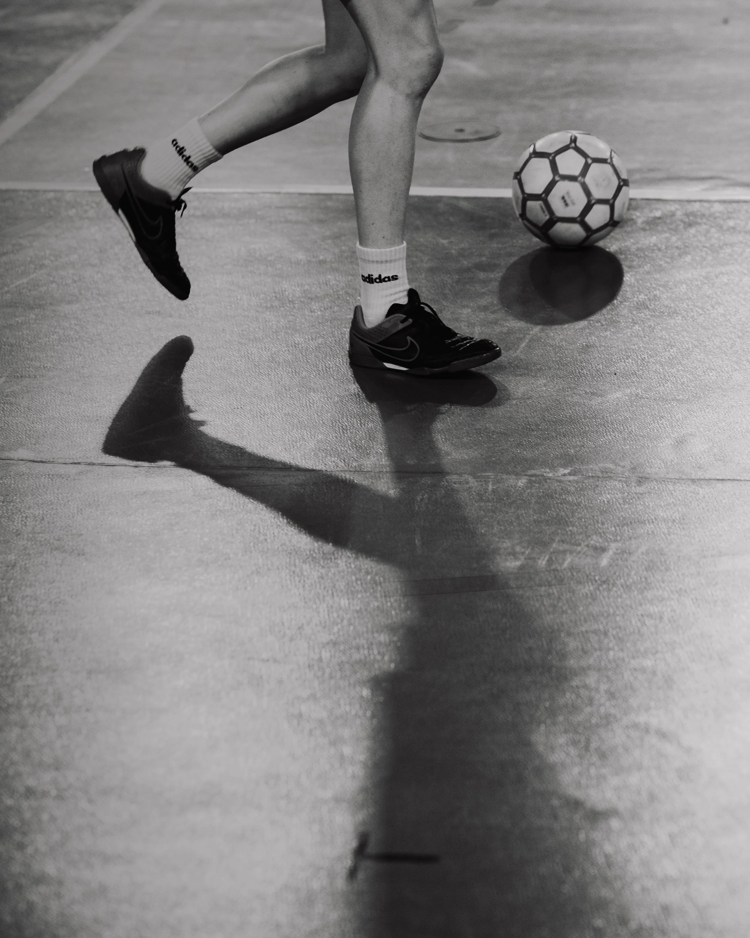 Un jeune joueur de football, en souliers de sport noirs, se déplace dans un gymnase, avec un ballon de football à proximité et une ombre projetée sur le sol.